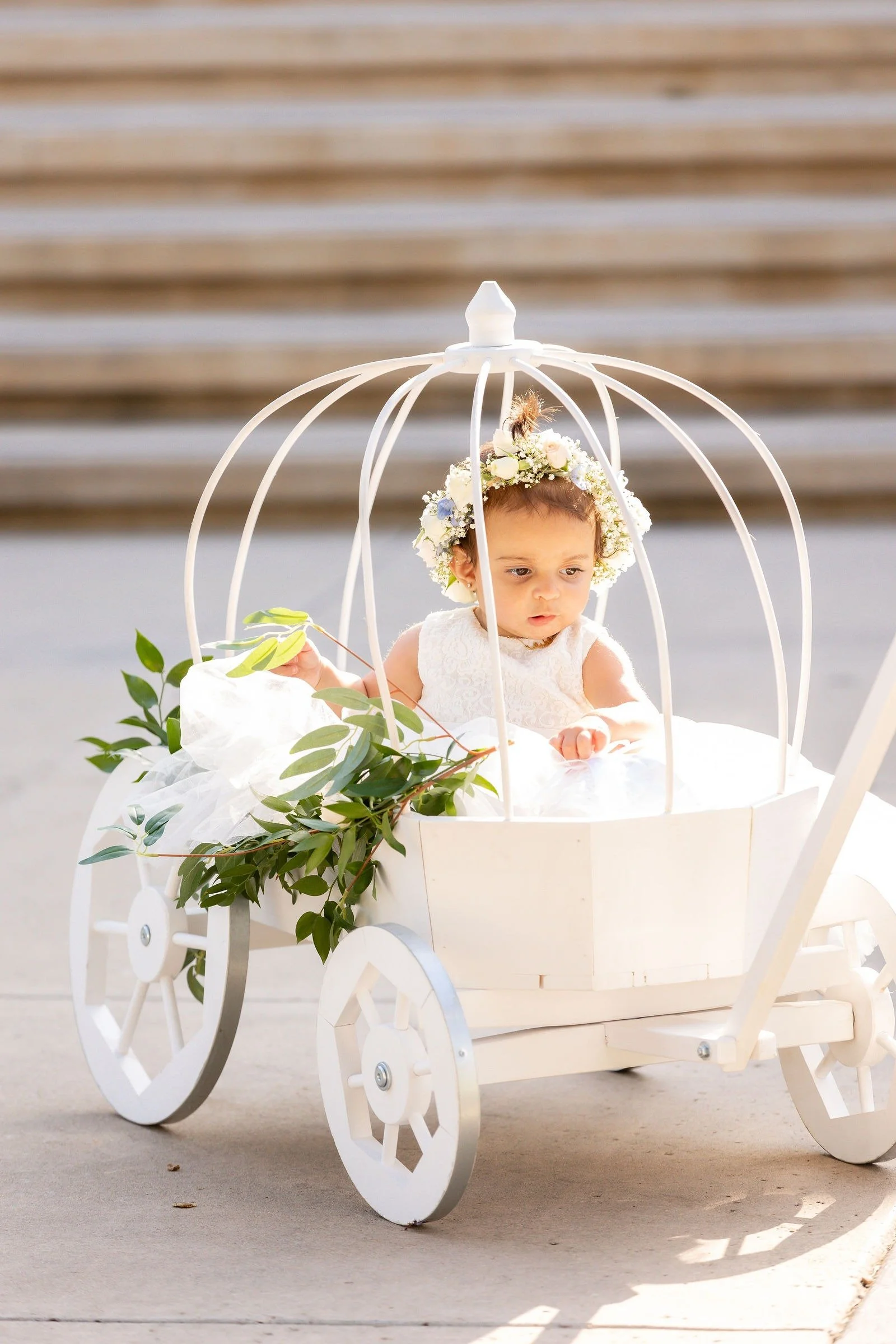 A young girl dressed in white, wearing a flower crown, sitting inside a small decorative white wagon with green leaves attached, on a sidewalk with steps in the background.