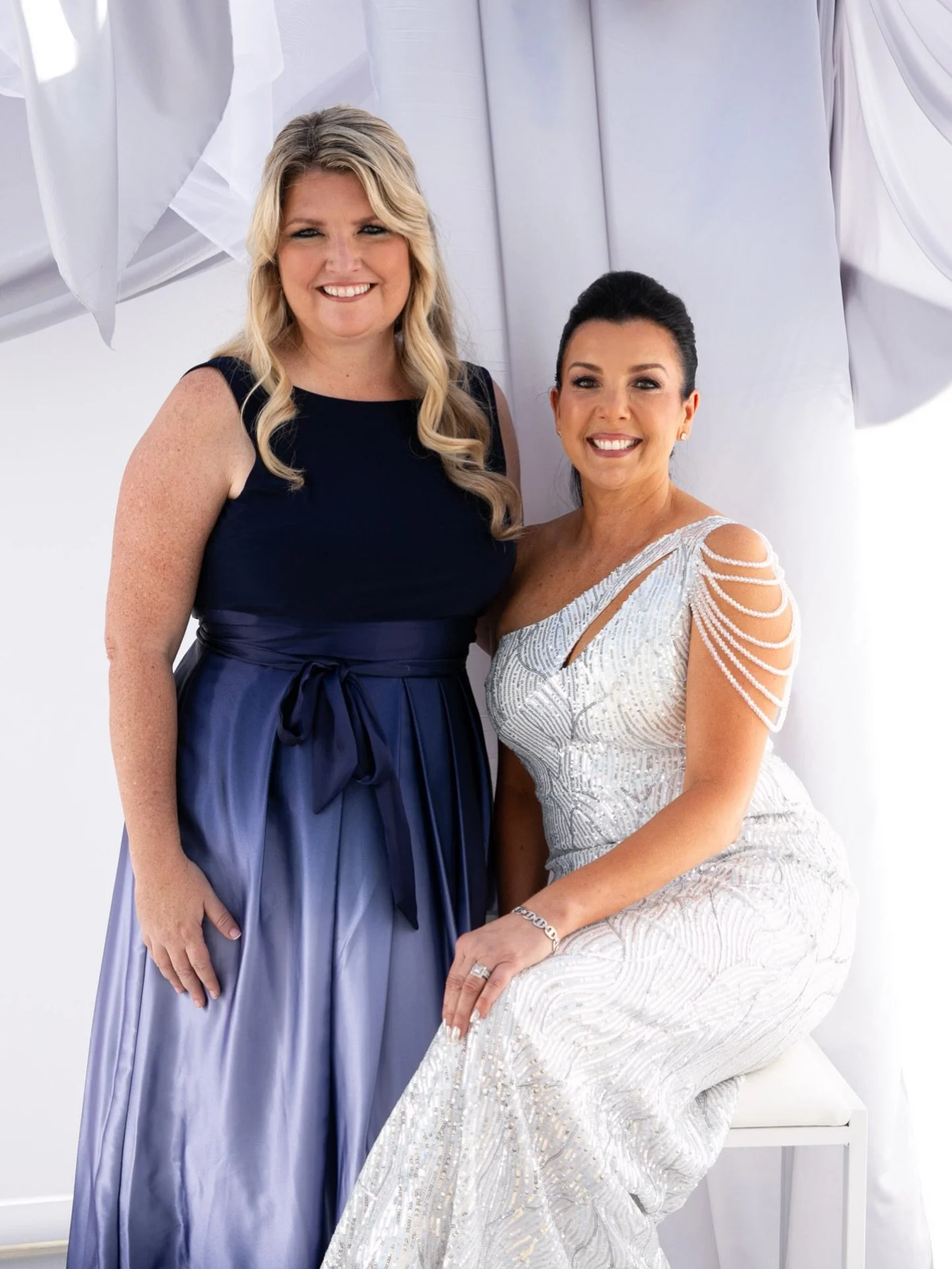 Two women dressed in elegant formal gowns pose together for a photo inside a white photo booth with draped fabric background, smiling.
