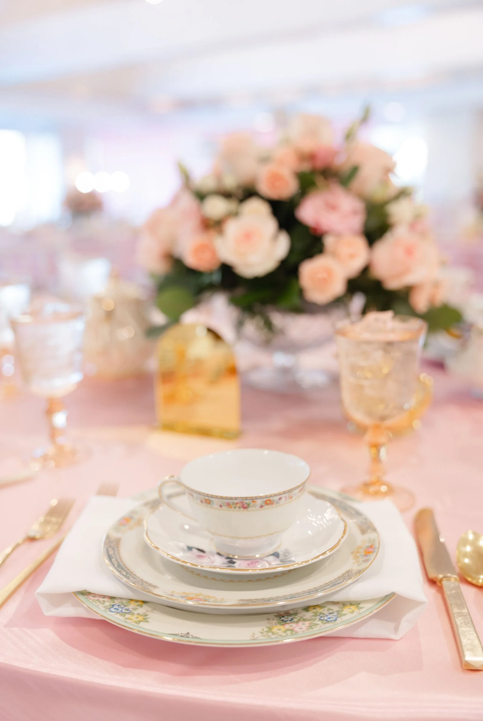 Elegant table setting with floral china, gold-accented teacup and saucer, set on a pink tablecloth, with pink and peach flowers in the background.