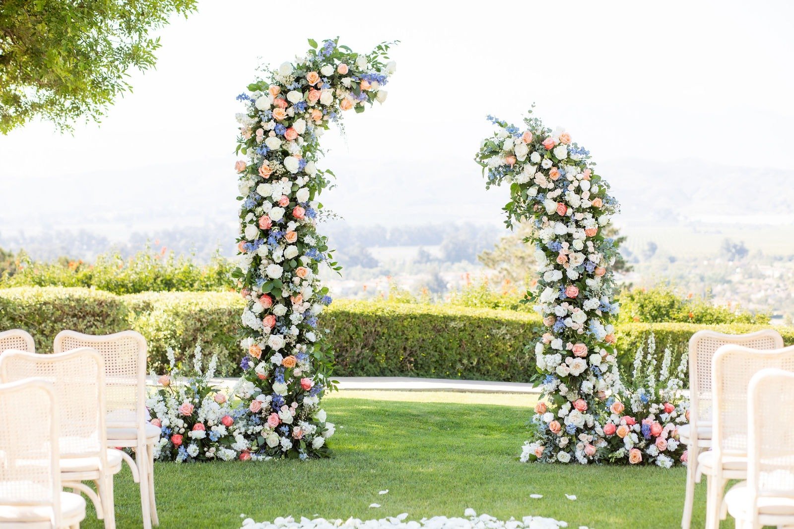 A wedding arch made of flowers, with white, pink, and blue blossoms, set up outdoors on a grassy area with chairs on either side, against a backdrop of greenery and a distant view.