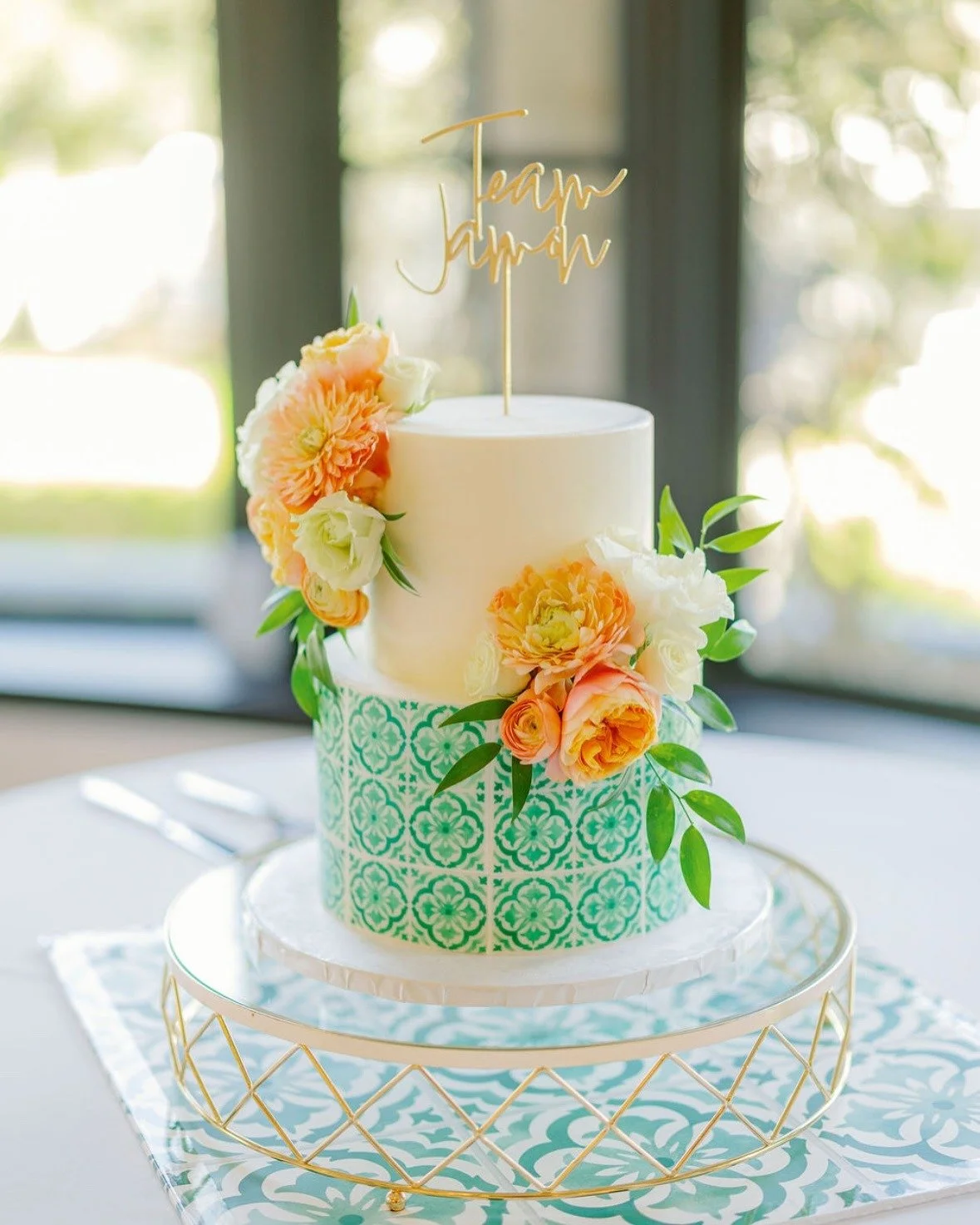 A two-tiered wedding cake decorated with peach and white flowers, with a gold 'Team Jovan' cake topper, sitting on a decorative gold stand near a window.