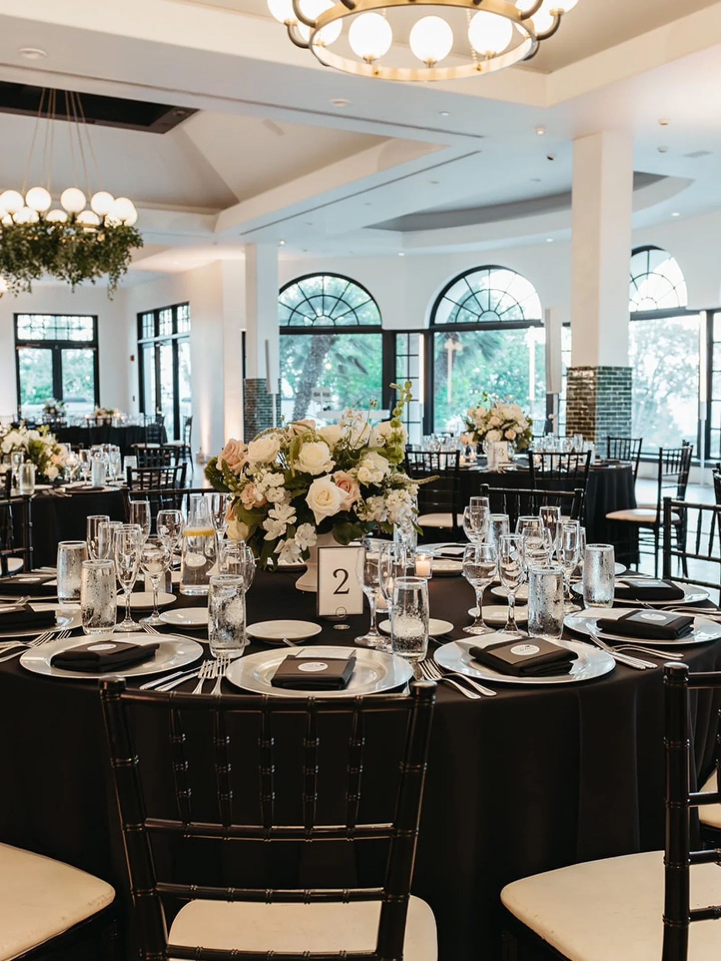 Round banquet table with black tablecloth, set with white plates, black napkins, silverware, champagne flutes, and water glasses, decorated with a floral centerpiece of white and blush roses, and greenery, in a bright event space with large windows a