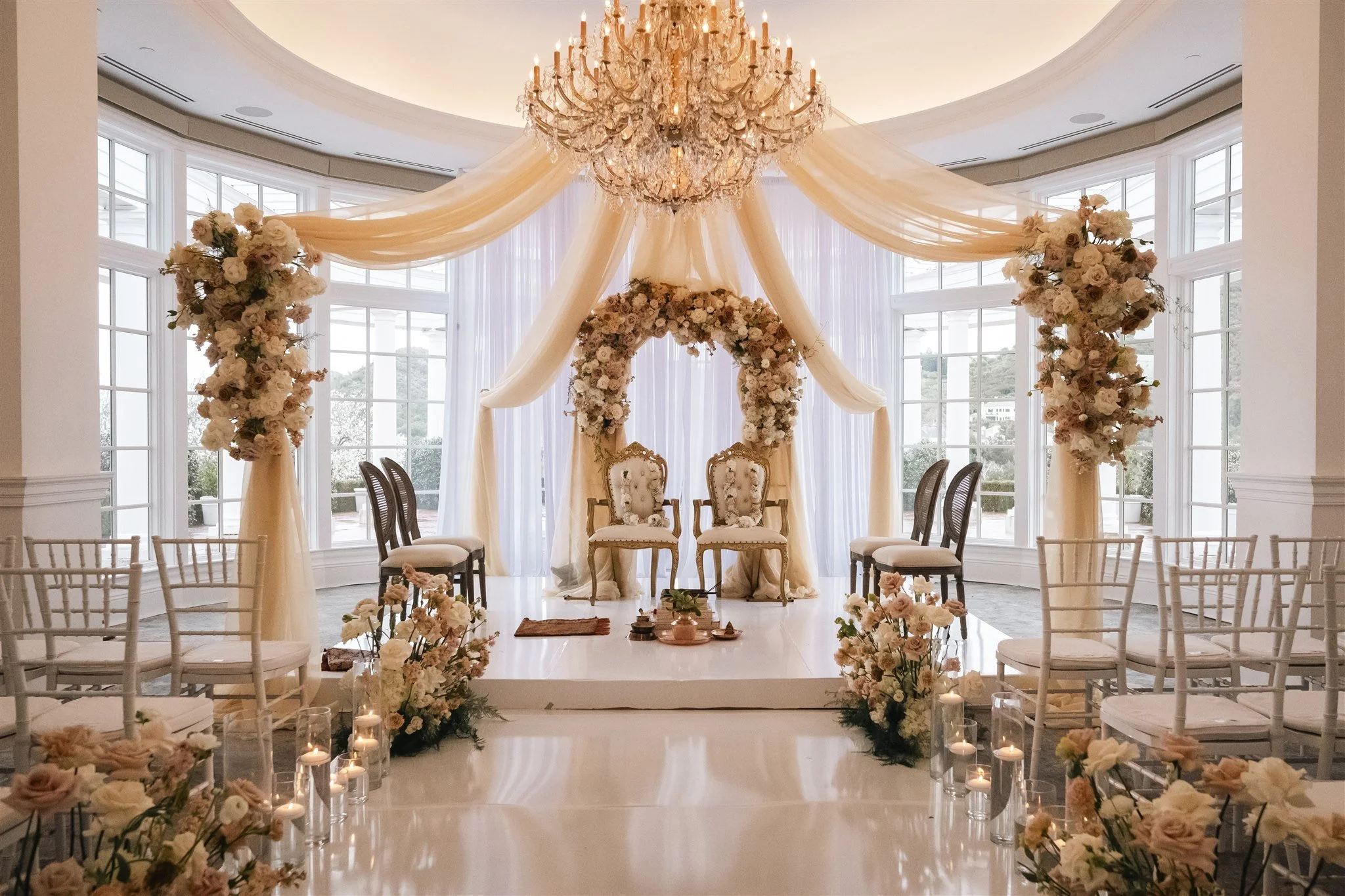 Elegant wedding ceremony setup inside a bright room with large windows, floral arrangements, a chandelier, and two ornate chairs under a floral arch.