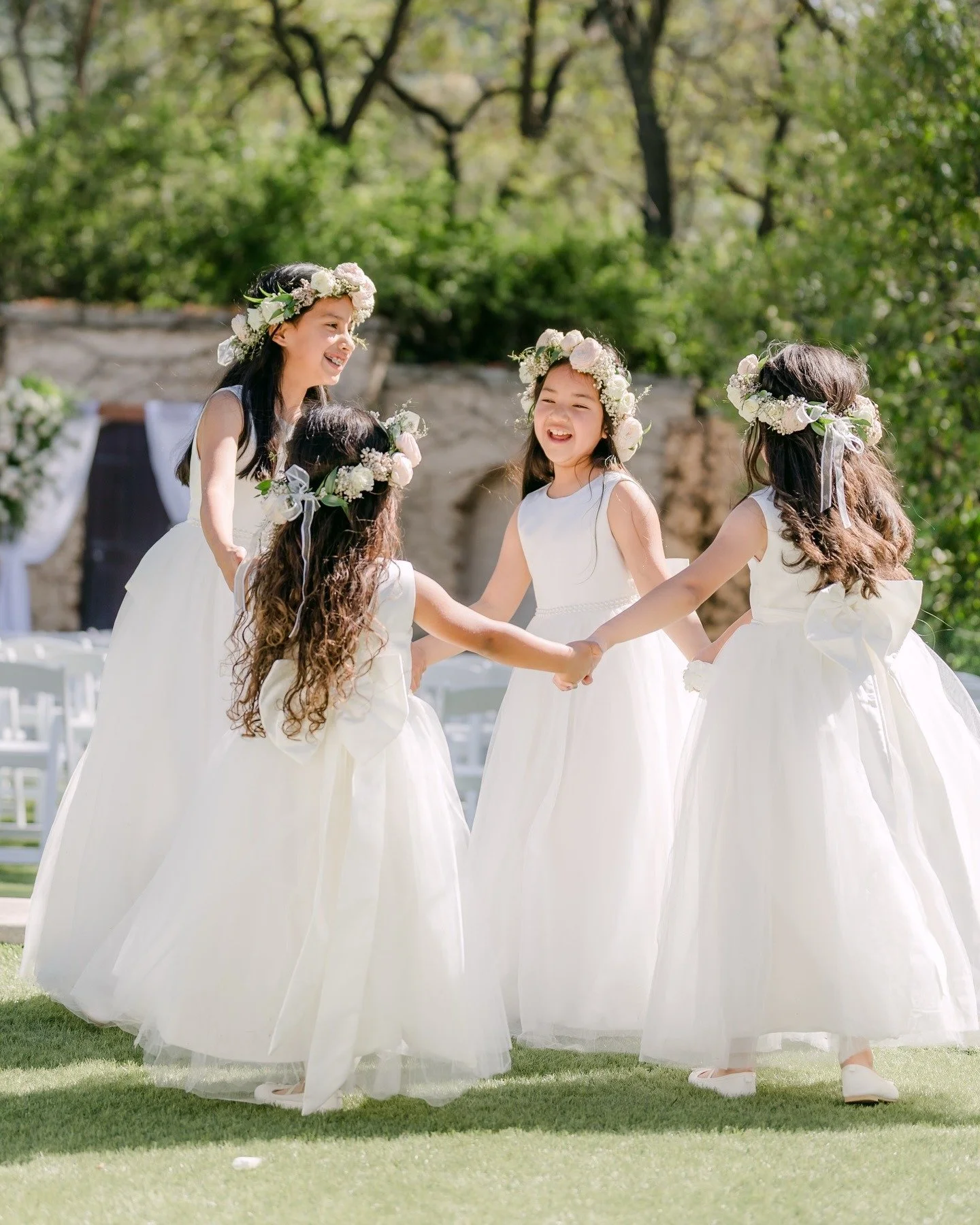 Five young girls wearing white dresses and floral crowns holding hands in a circle outdoors, smiling and playing together on a sunny day.