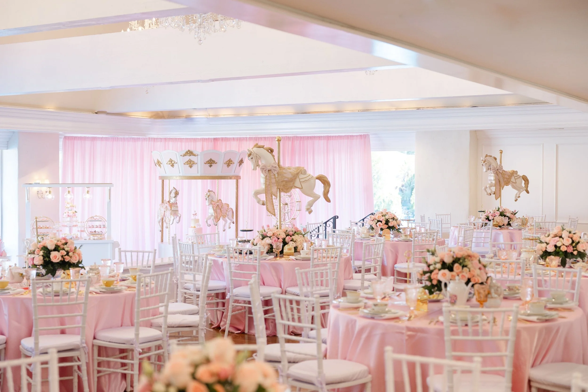 Elegant wedding reception with round tables dressed in pink tablecloths, adorned with pink and white flower centerpieces, and decorated with carousel horse and horse figurine centerpieces. The room features white chairs, soft pink curtains, and a chandelier.