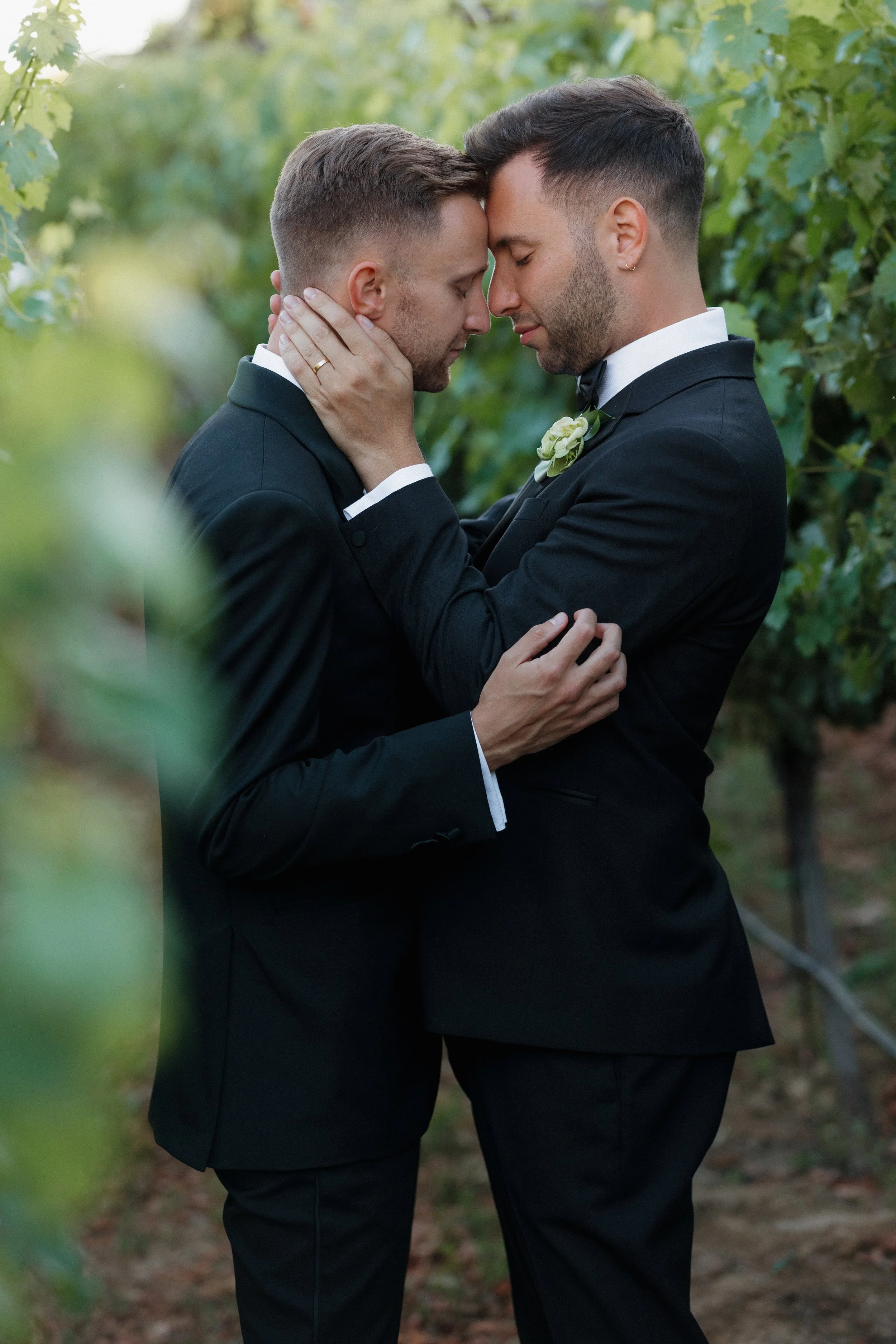 Two men in tuxedos embracing each other in a vineyard.