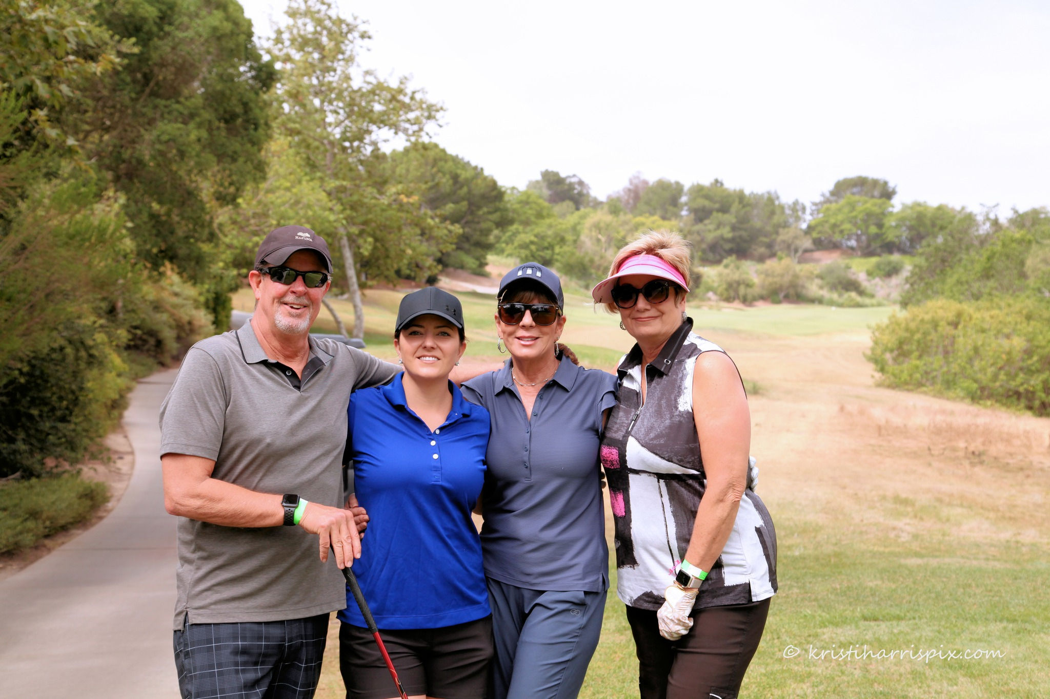 Group of four people standing outdoors on a golf course, smiling and looking at the camera, with trees and grass in the background.