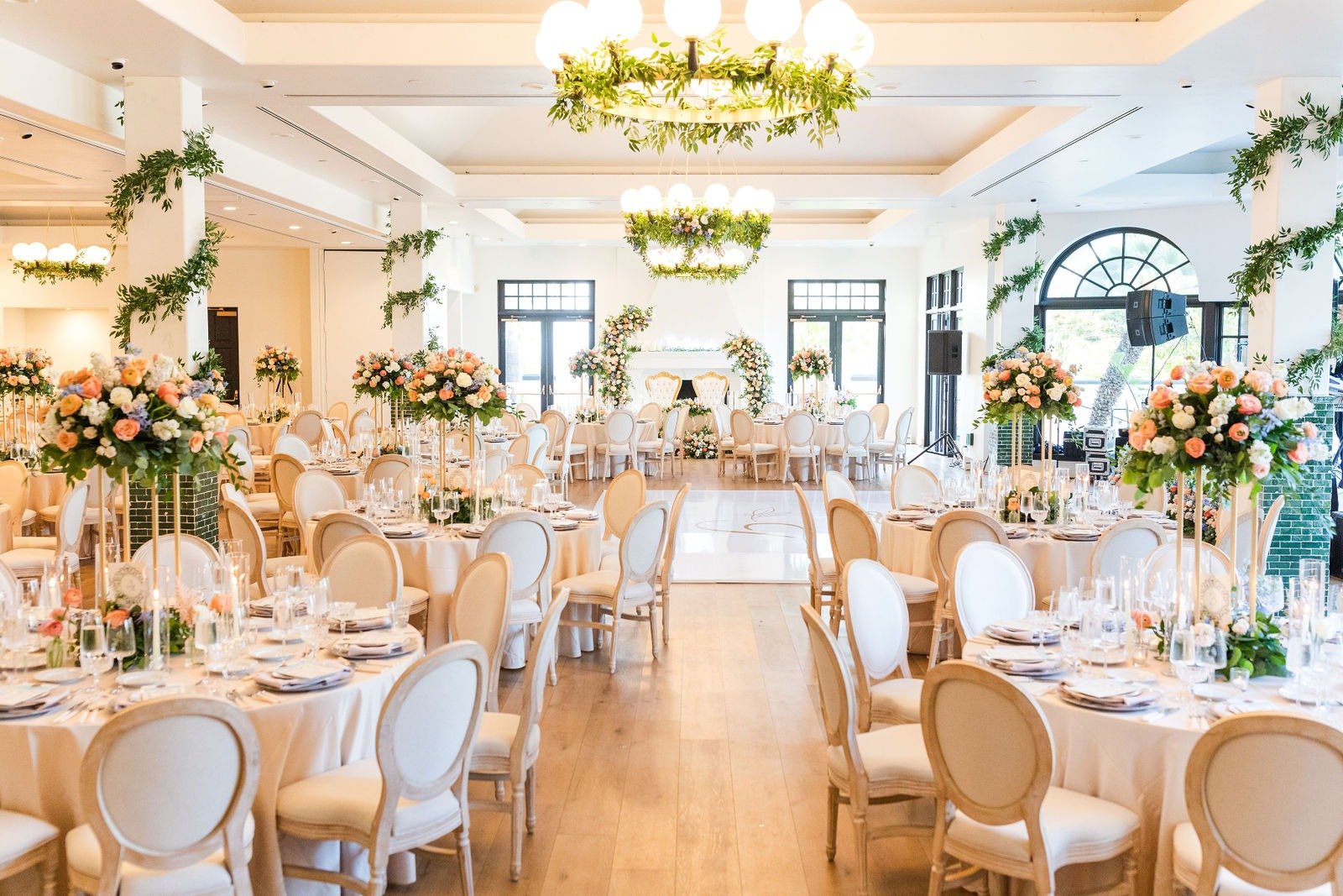Elegant banquet hall decorated for wedding reception with round tables, cream-colored chairs, floral centerpieces, and an arch at the head table, with large windows and greenery accents.