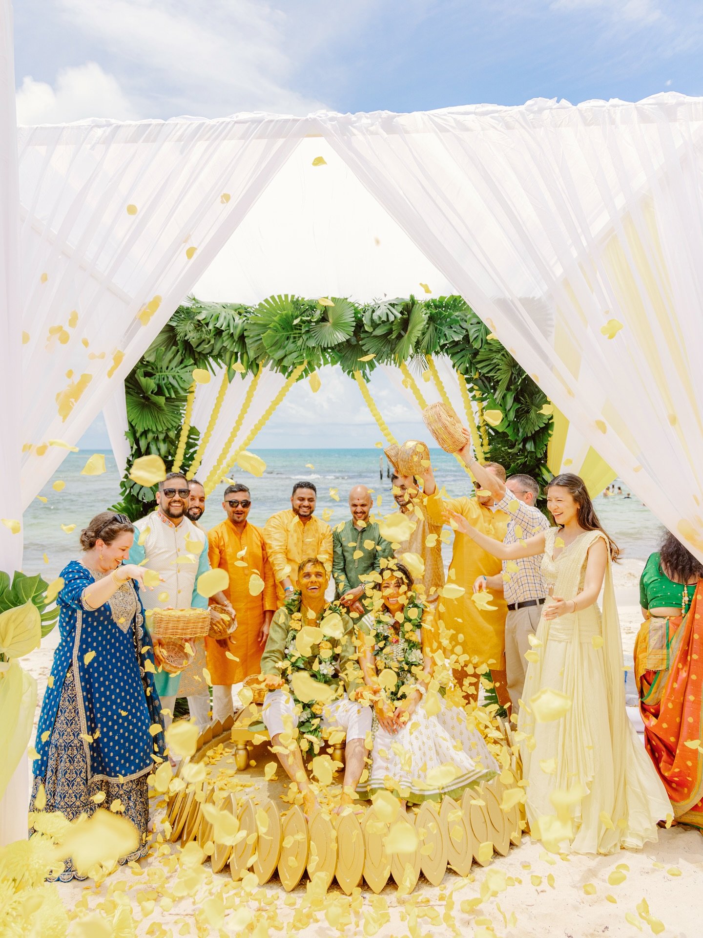 A beach wedding ceremony with a group of people celebrating under a decorated canopy, with yellow flower petals in the air and ocean in the background.
