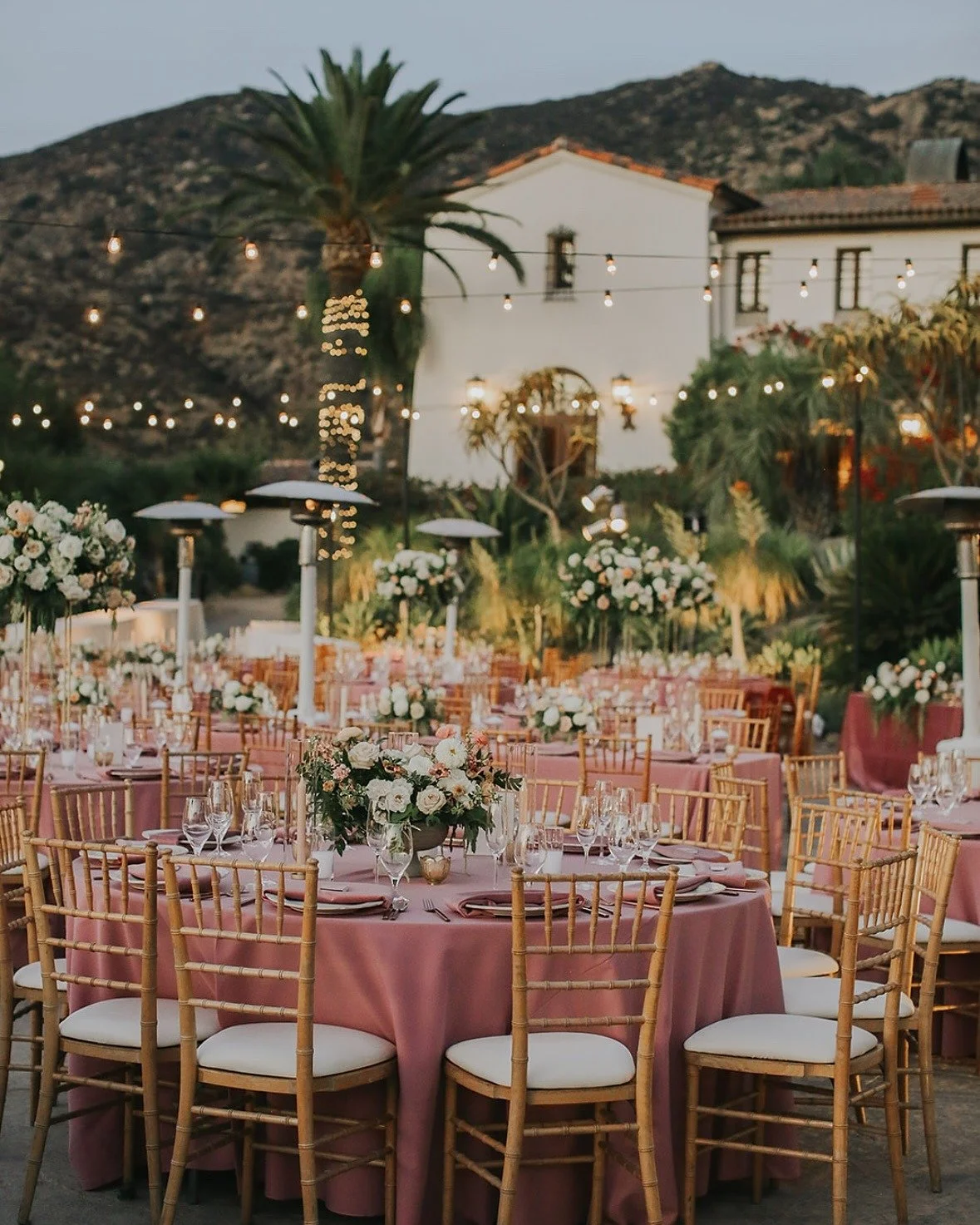 Outdoor wedding reception setup with round tables covered in pink tablecloths, adorned with floral centerpieces and surrounded by wooden chairs with white cushions. String lights are hanging overhead, and heaters are in place for warmth. In the backg
