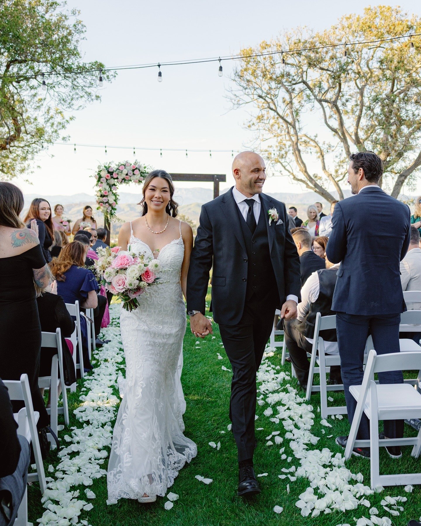 Bride and groom walking down the aisle at their outdoor wedding ceremony, smiling and holding hands, surrounded by guests and decorated with flower petals and a floral arch.