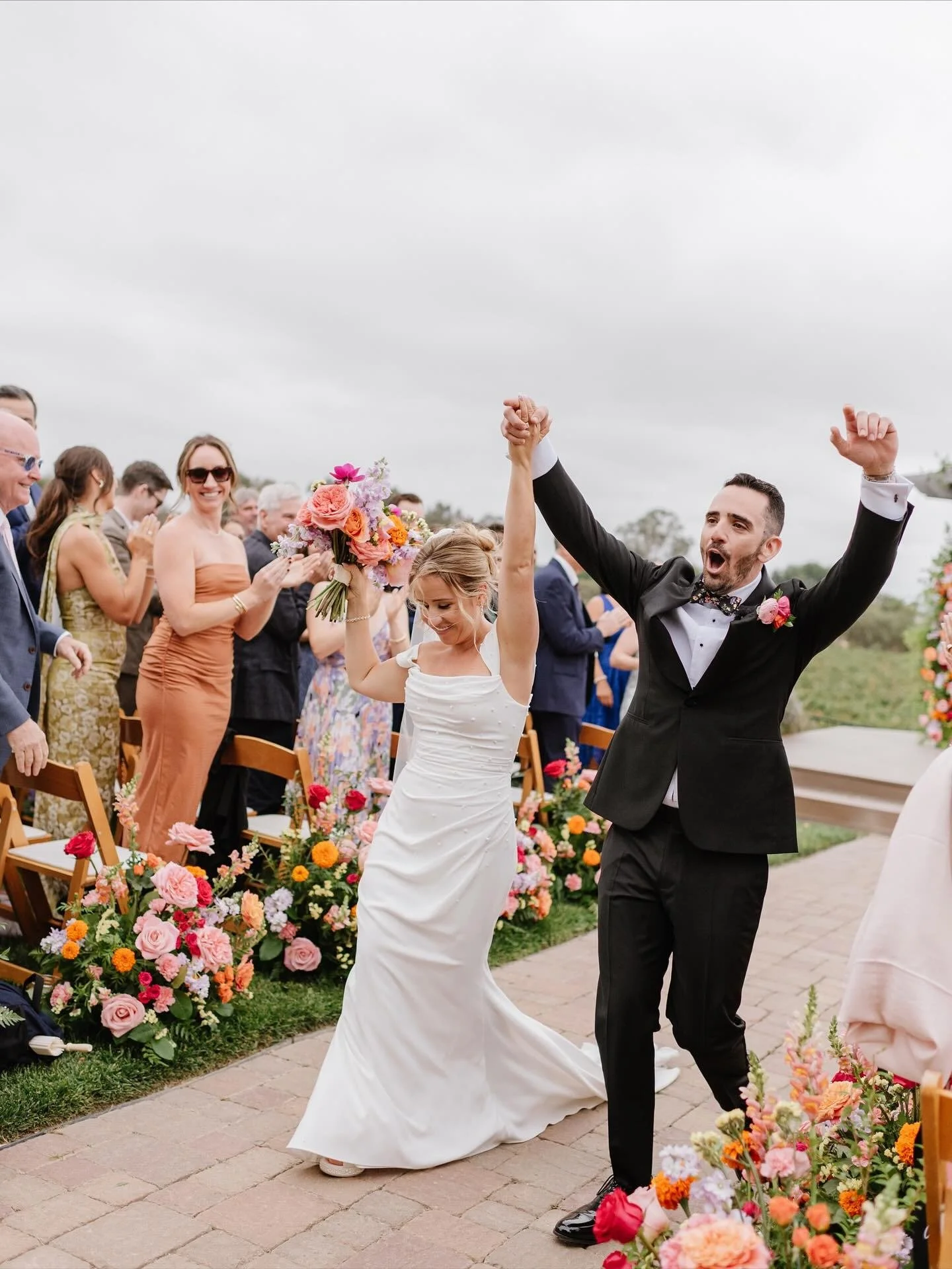 A newlywed couple celebrating their wedding outdoors, the bride holding a bouquet, and the groom raising his fist in victory, as guests cheer around them, with floral arrangements lining the aisle.