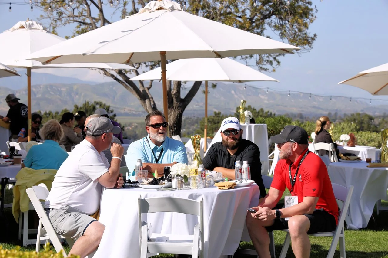 Group of people sitting around a table outdoors under white umbrellas, enjoying a event with scenic mountain views in the background.