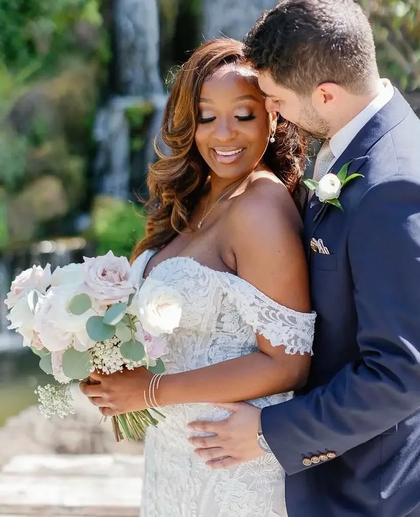 A bride and groom embrace outdoors during their wedding, the bride holding a bouquet of white and pink flowers, the bride wearing a lace off-shoulder wedding dress, and the groom in a navy suit with a white boutonniere.