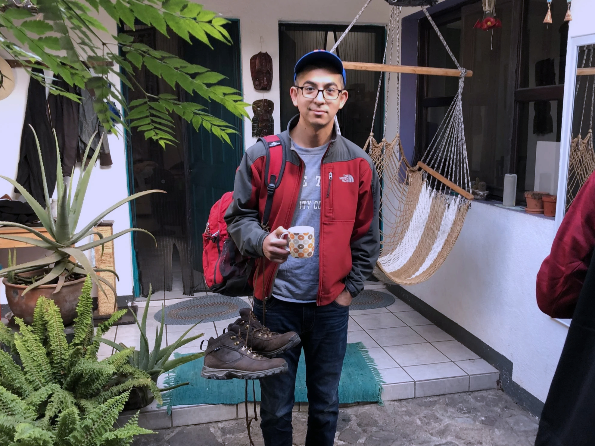 A young man wearing glasses, a red and gray jacket, and a blue cap, holding a mug of coffee with a colorful pattern, standing on a tiled porch with a hammock hanging in the background, surrounded by plants.