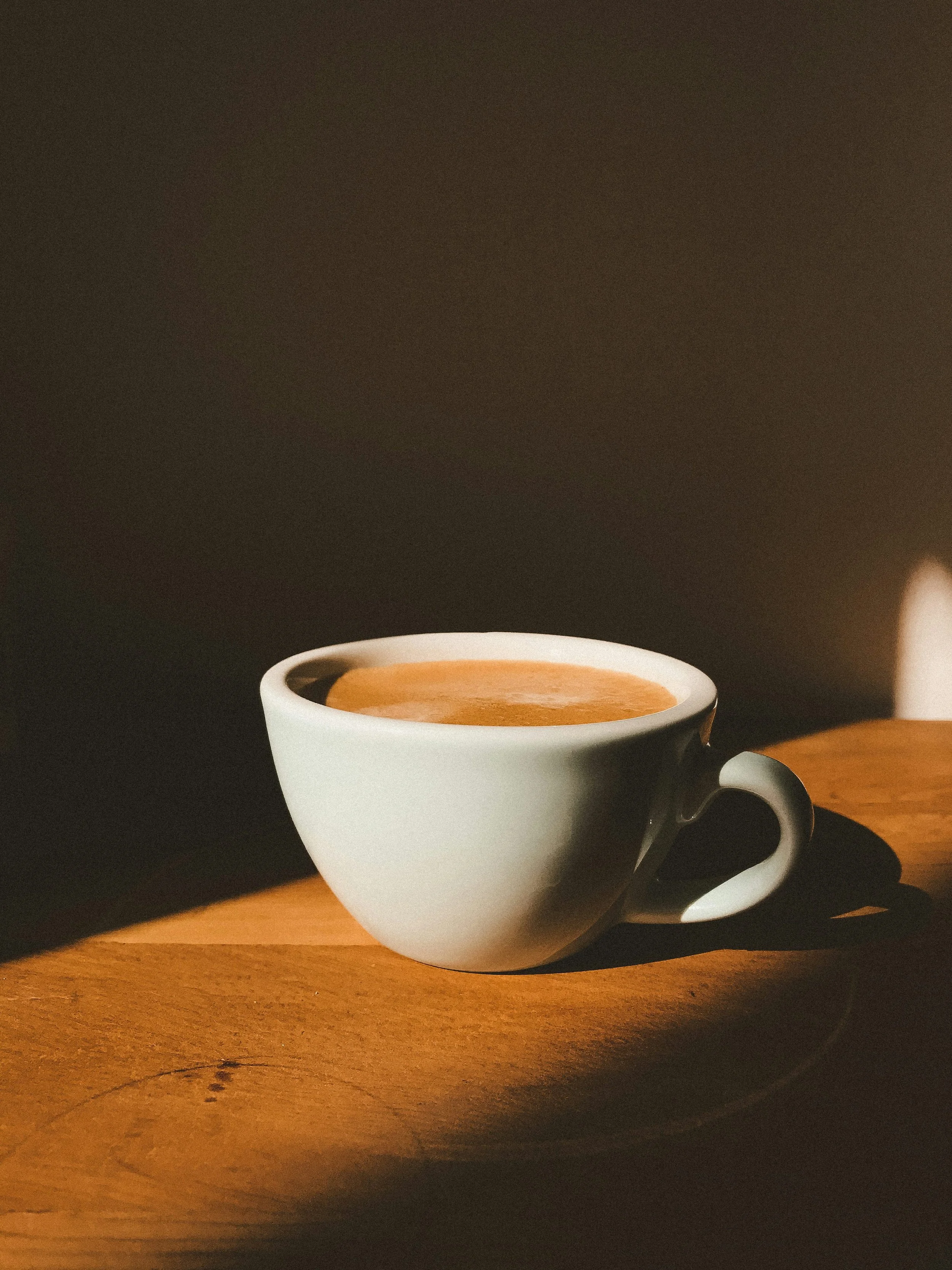 White coffee cup filled with coffee on a wooden surface, with sunlight creating shadows.