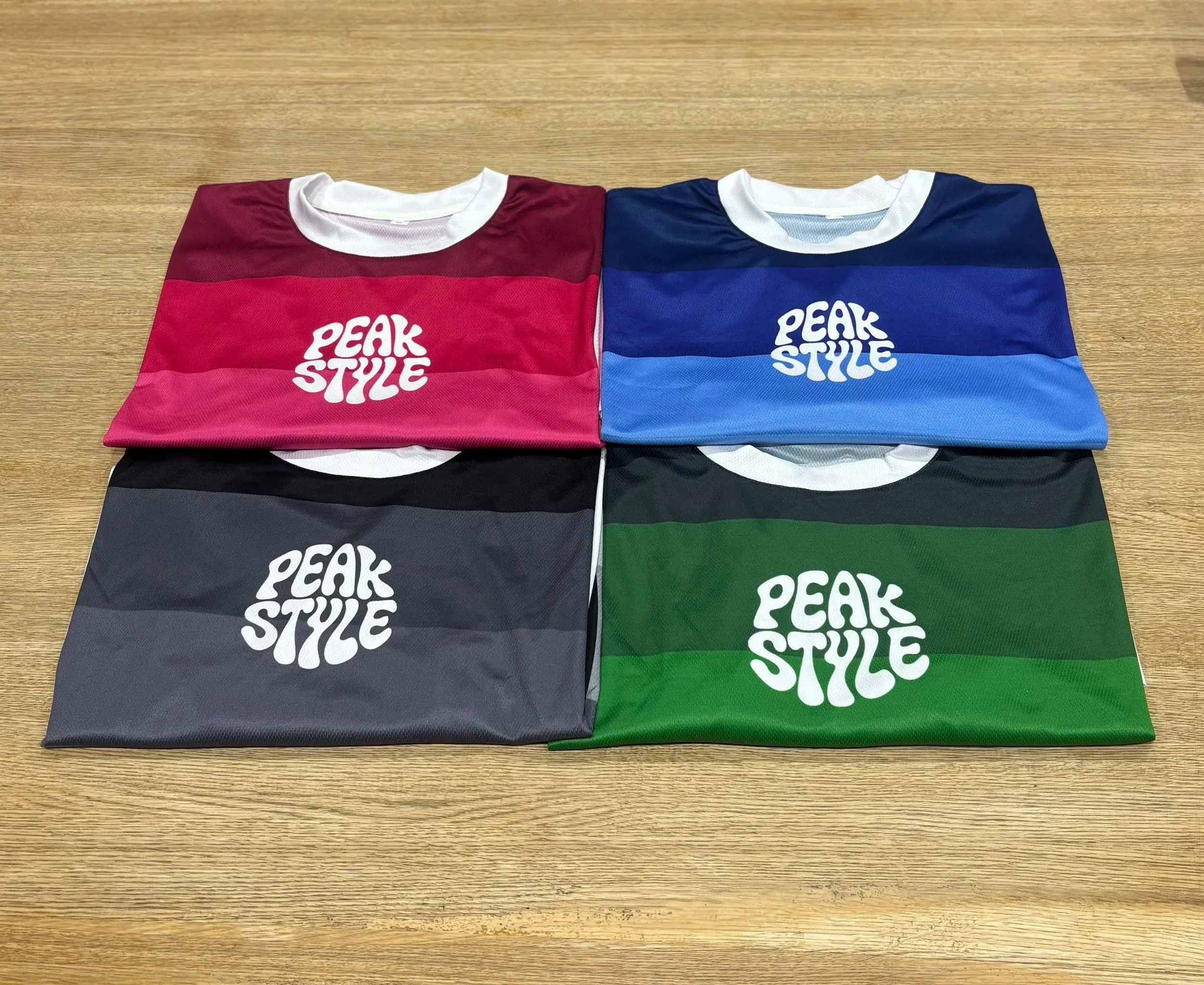 Four folded short-sleeve shirts on a wooden surface, each with a different color combination and the white 'PEAK STYLE' logo on the front.