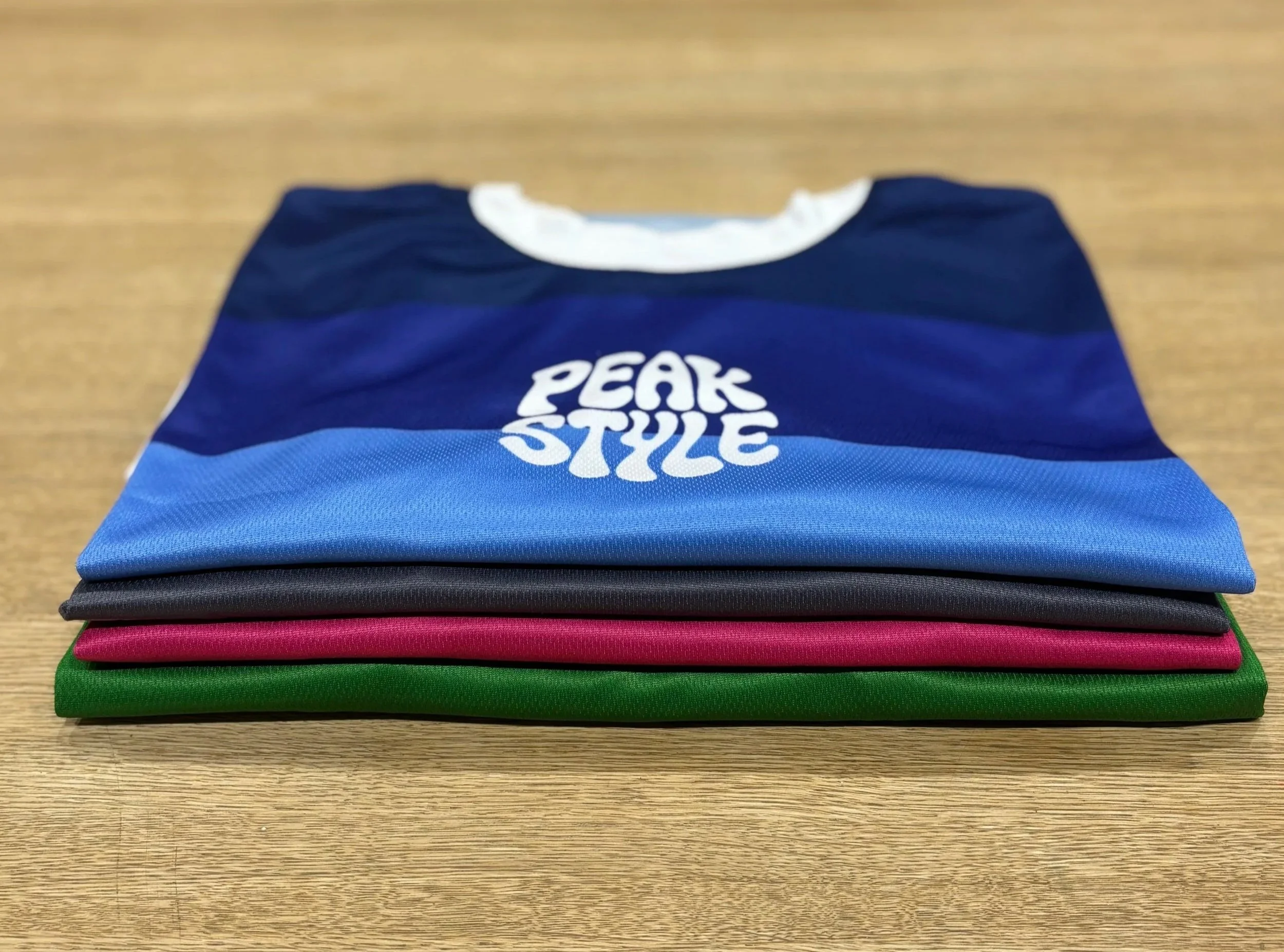 Folded colorful athletic shirts on a wooden surface, one with 'PEAK STYLE' printed on it.
