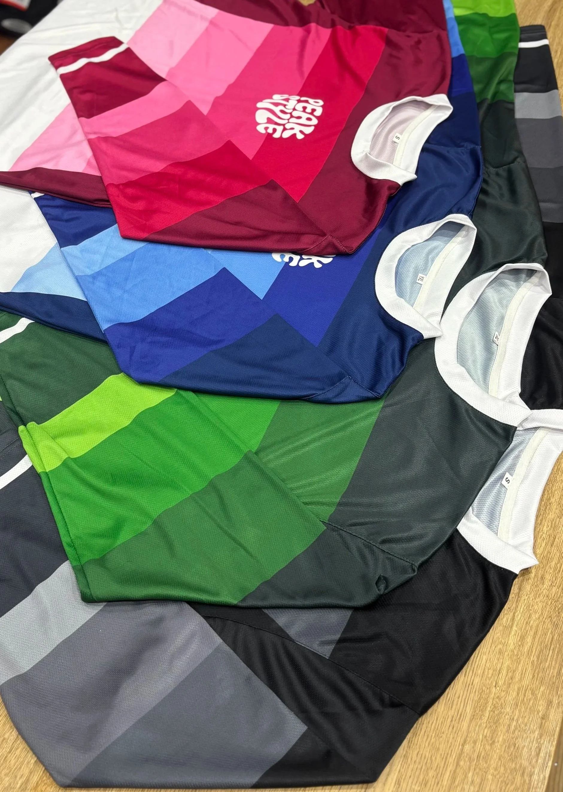 Multiple colorful sports jerseys arranged in a row on a wooden surface, including red, blue, green, black, and gray, with some showing white inside the collar area.