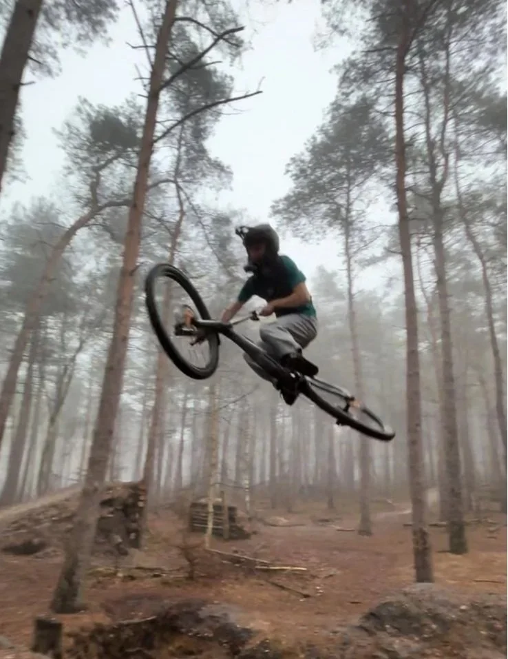 A person riding a mountain bike is mid-air in a forest with tall trees and dirt terrain.