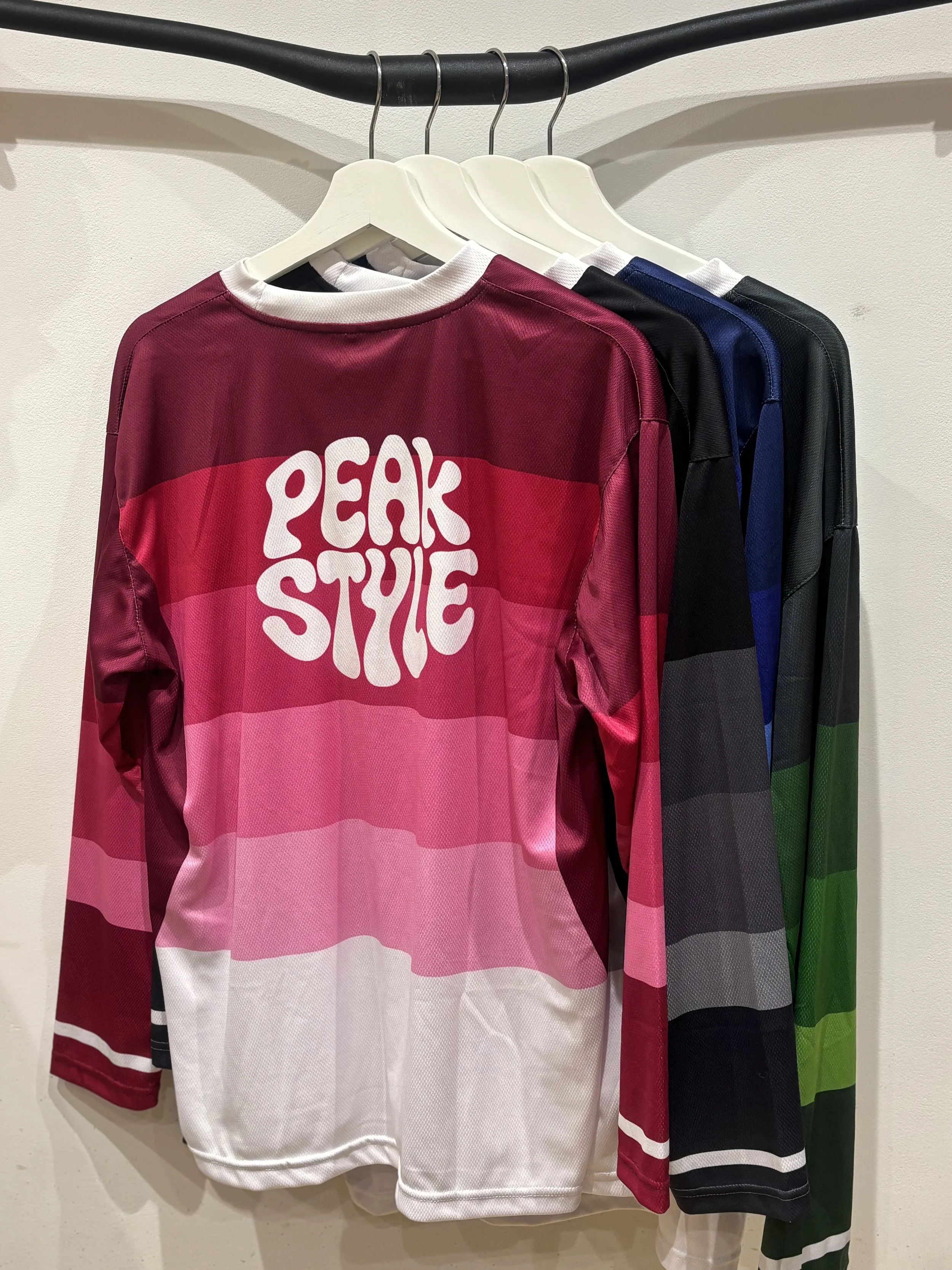 Four sports jerseys hanging on a rack with a white background, featuring a gradient of colors and the text 'PEAK STYLE' on the front of the first jersey.
