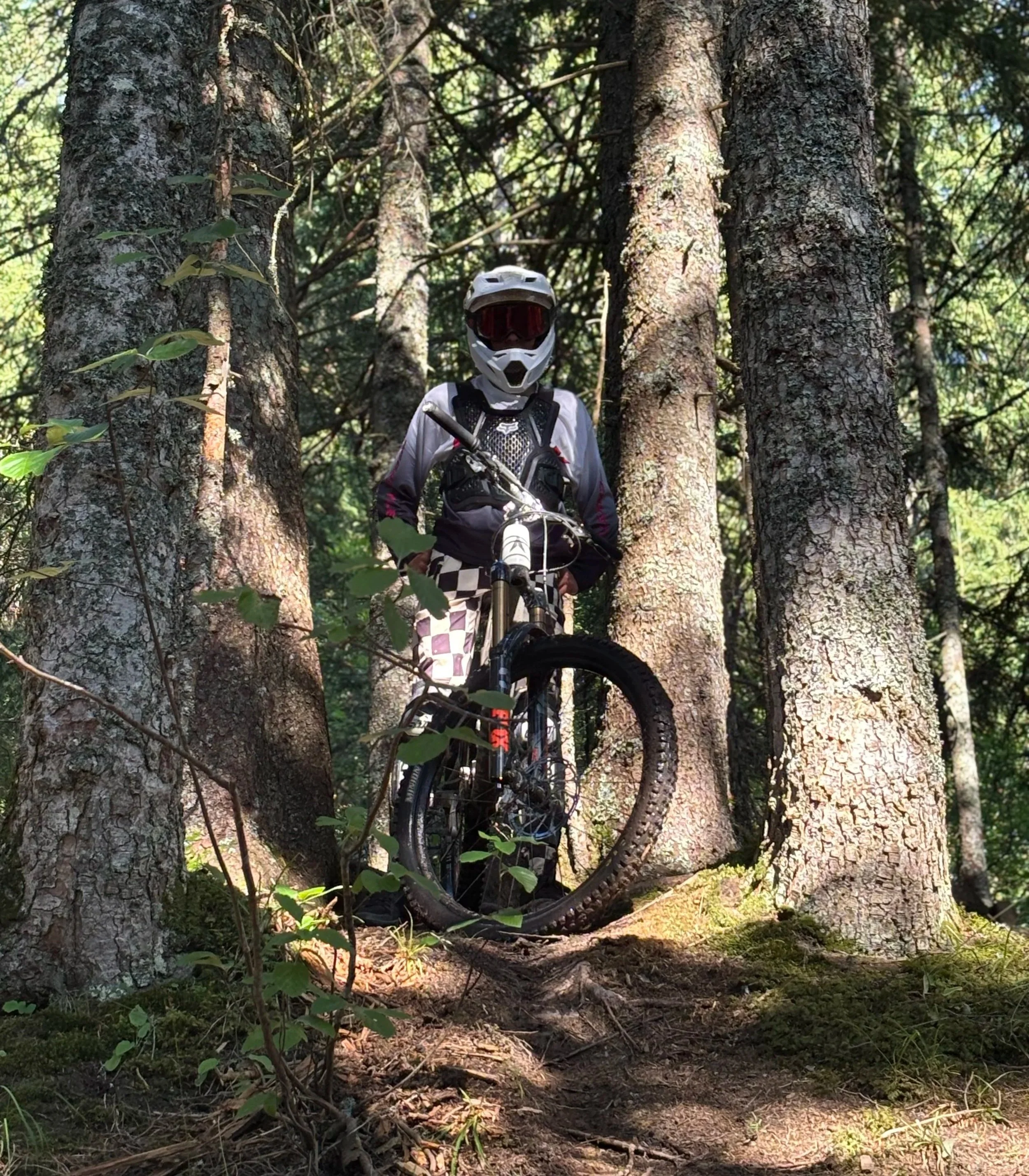 Person in full mountain biking gear standing with mountain bike on trail in forest, wearing helmet and protective clothing.