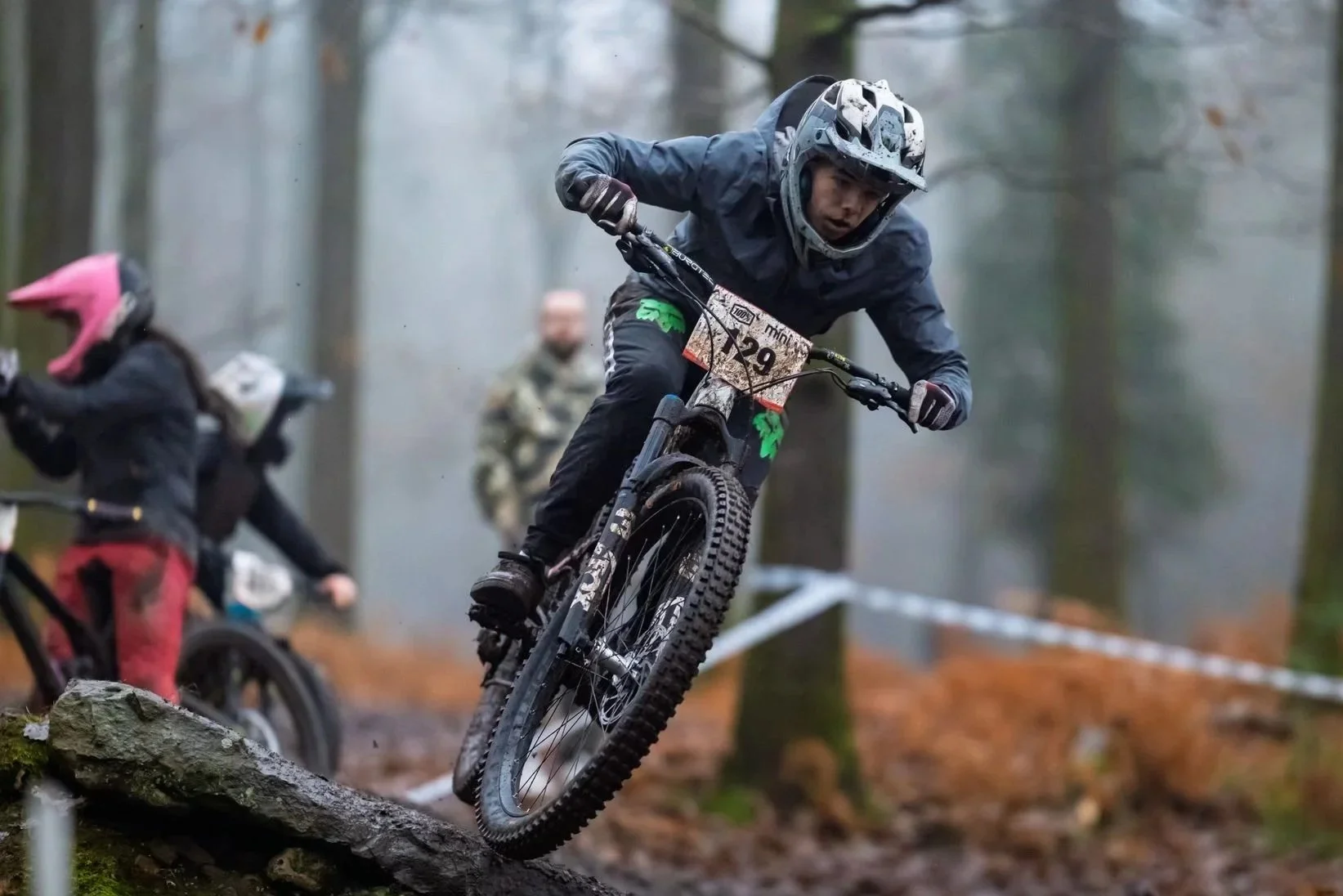 A person riding a mountain bike downhill on a forest trail during a race, wearing a helmet and protective gear, with other riders in the background.