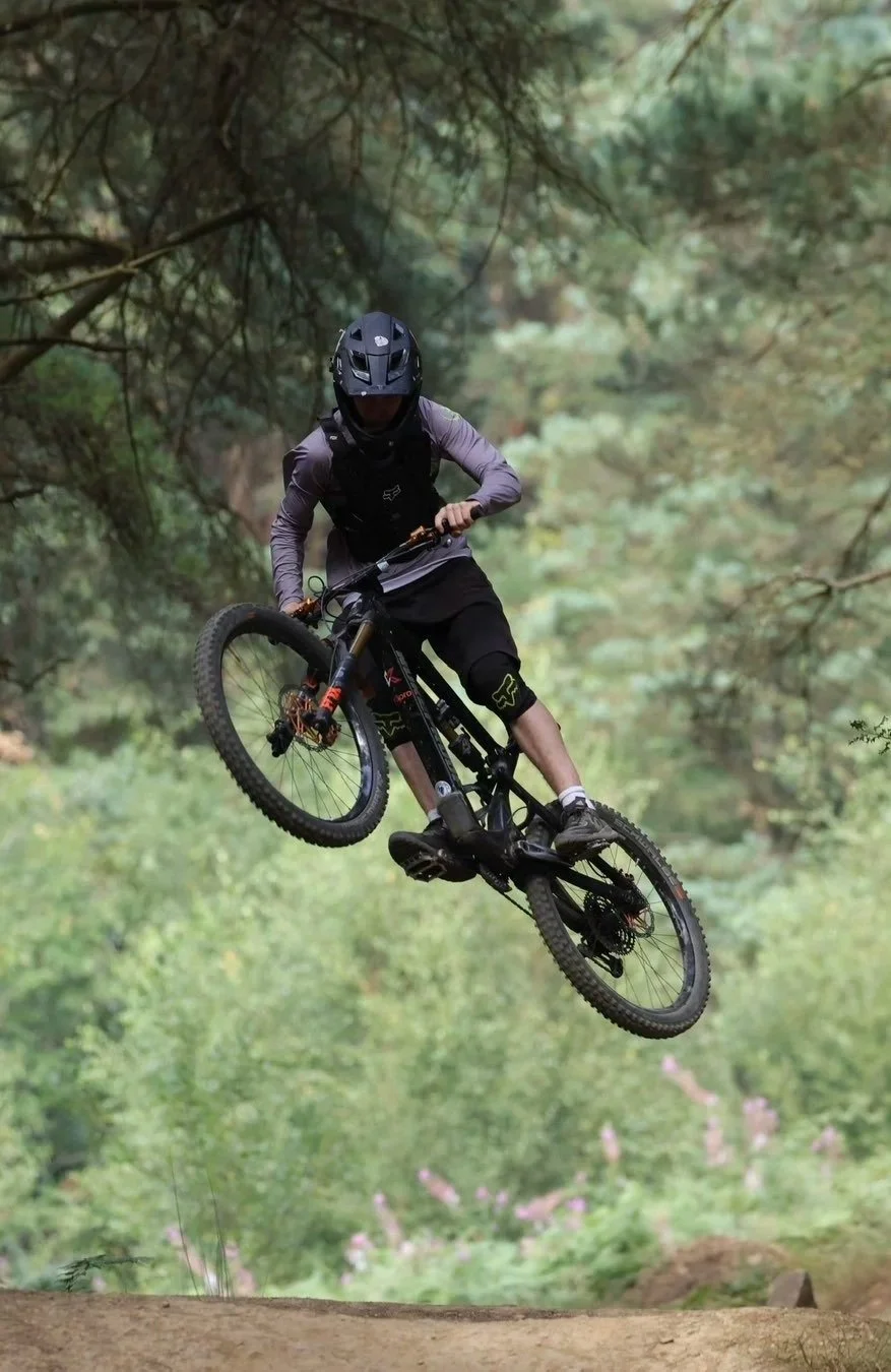 A mountain biker in mid-air performing a jump on a trail surrounded by trees.