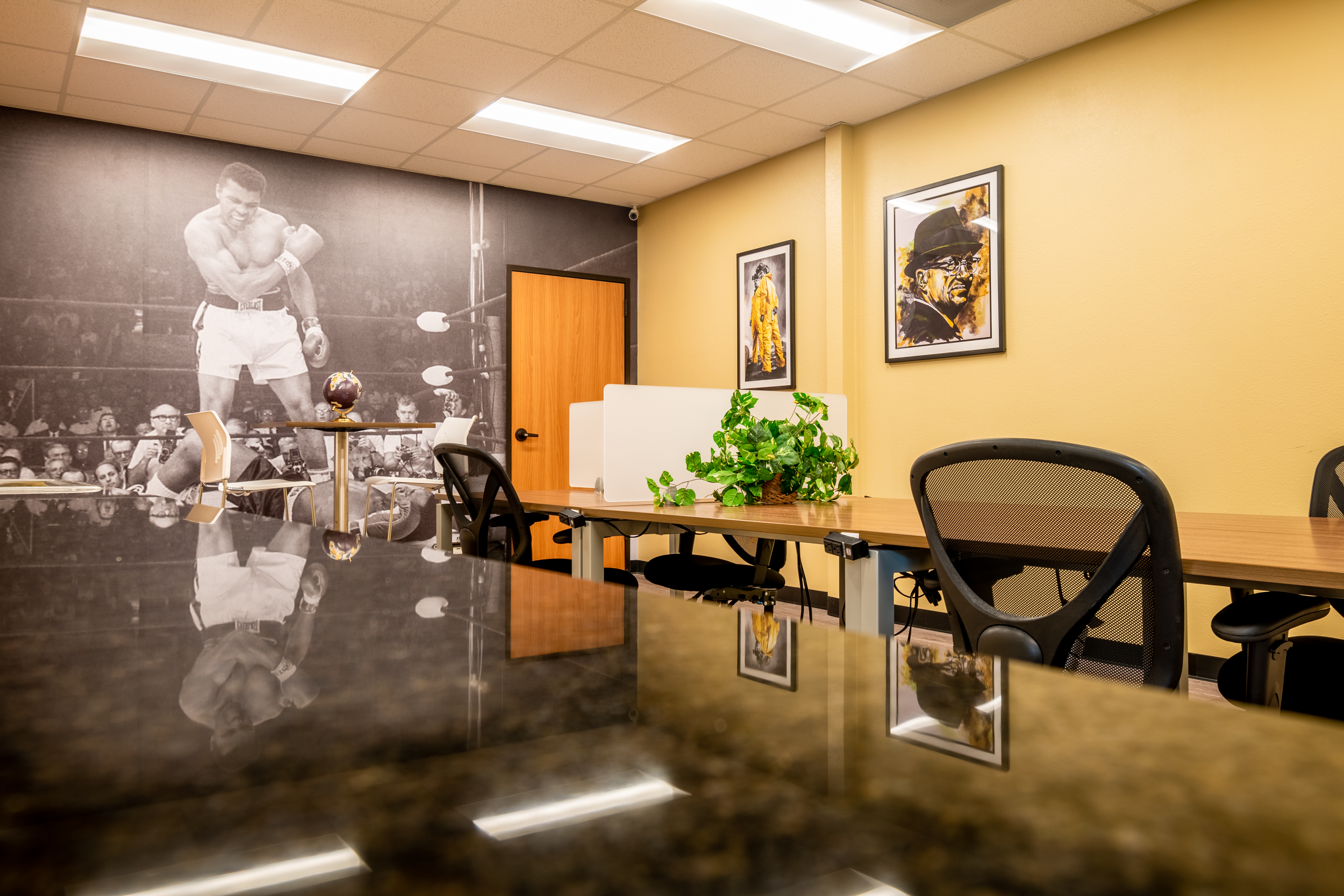 An office conference room with yellow walls, framed artwork, a large black-and-white photograph of a boxing match, a wooden table with office chairs, a green plant, and a door in the background.