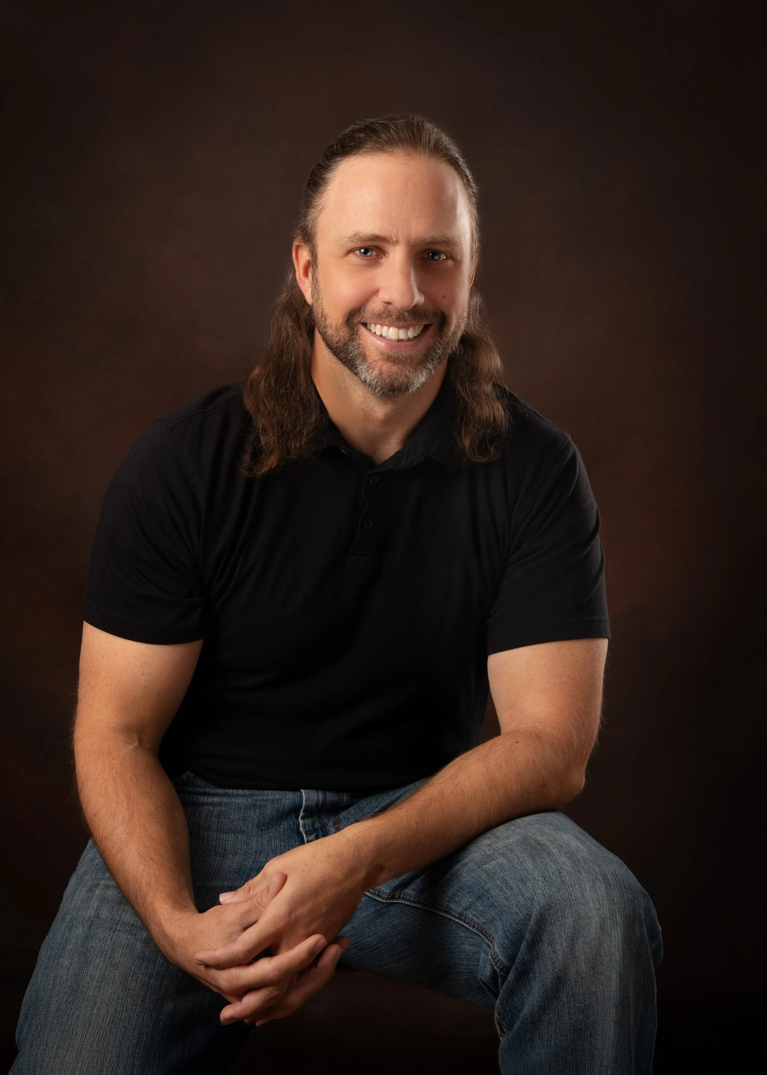 A man with long brown hair, a beard, and light skin, sitting and smiling, wearing a black shirt and jeans, against a dark brown background.