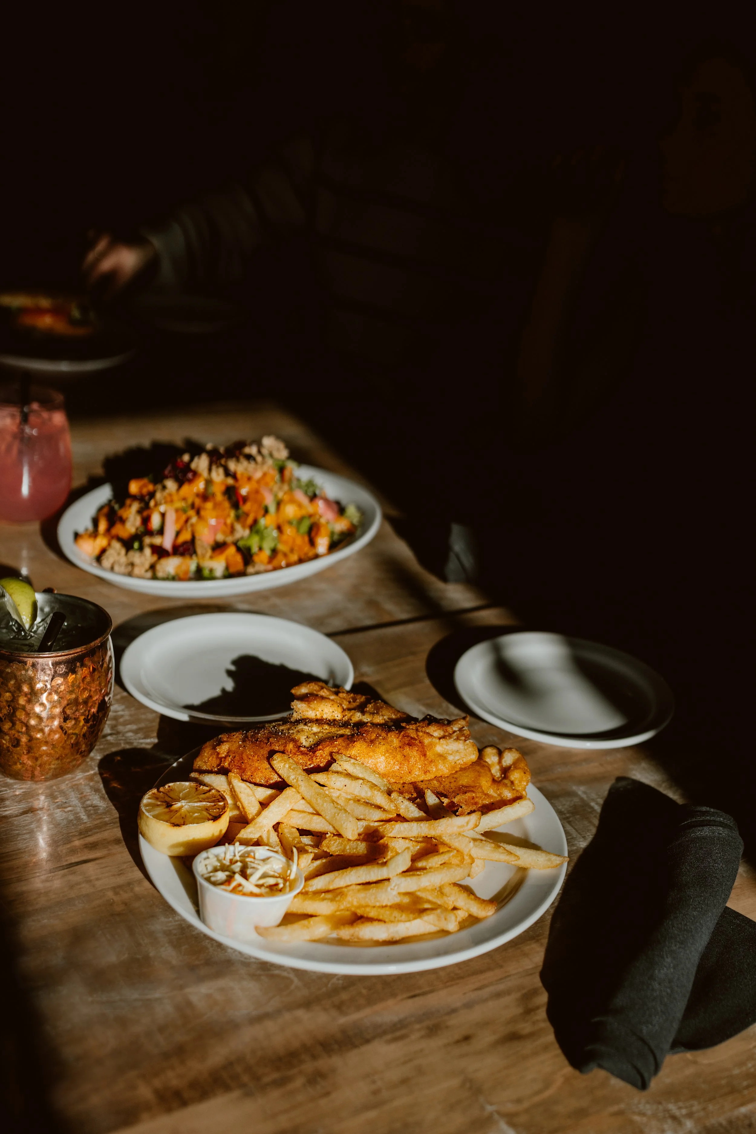 A plate of delicious  fish and chips with a lemon wedge and tartar sauce, a side of salad, and drinks on a wooden table at The Joyce Pub.