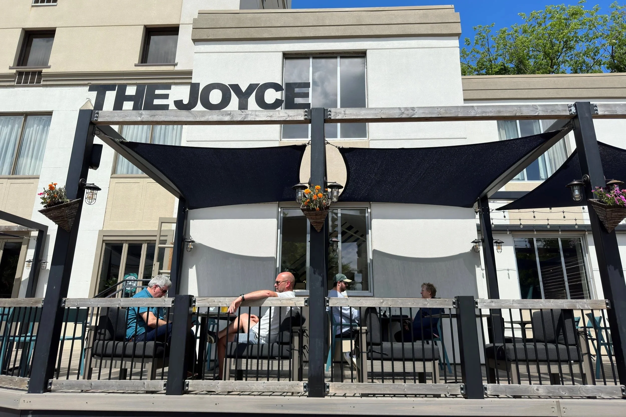 Outdoor patio of a building named THE JOYCE with people sitting at tables and a black shade structure, potted plants hanging on posts, and the building of the Crowne Plaza Fredericton- Lord Beaverbrook Hotel in the background.