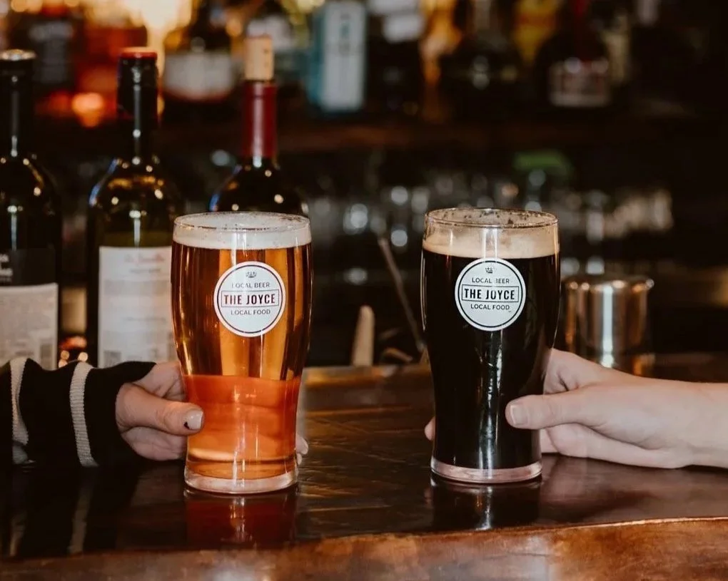 Two glasses of craft beer being held by people at a bar, with a background of liquor bottles. One glass contains amber beer, and the other is dark stout. Both glasses have labels reading 'The Joyce, Local Beer, Local Food.'