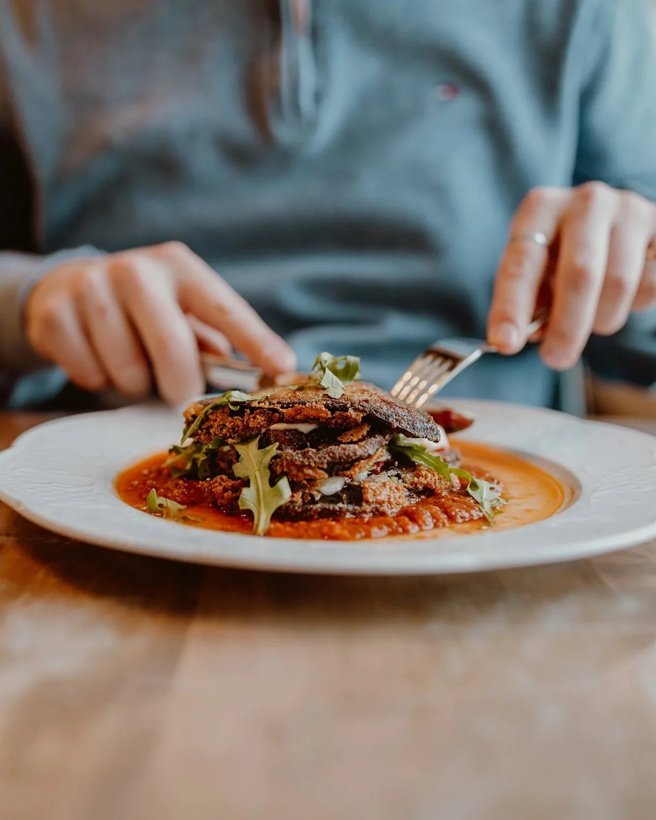 Person eating a plate of food, featuring a stack of fried eggplant with greens on top, served on a white plate with sauce underneath at The Joyce Pub.