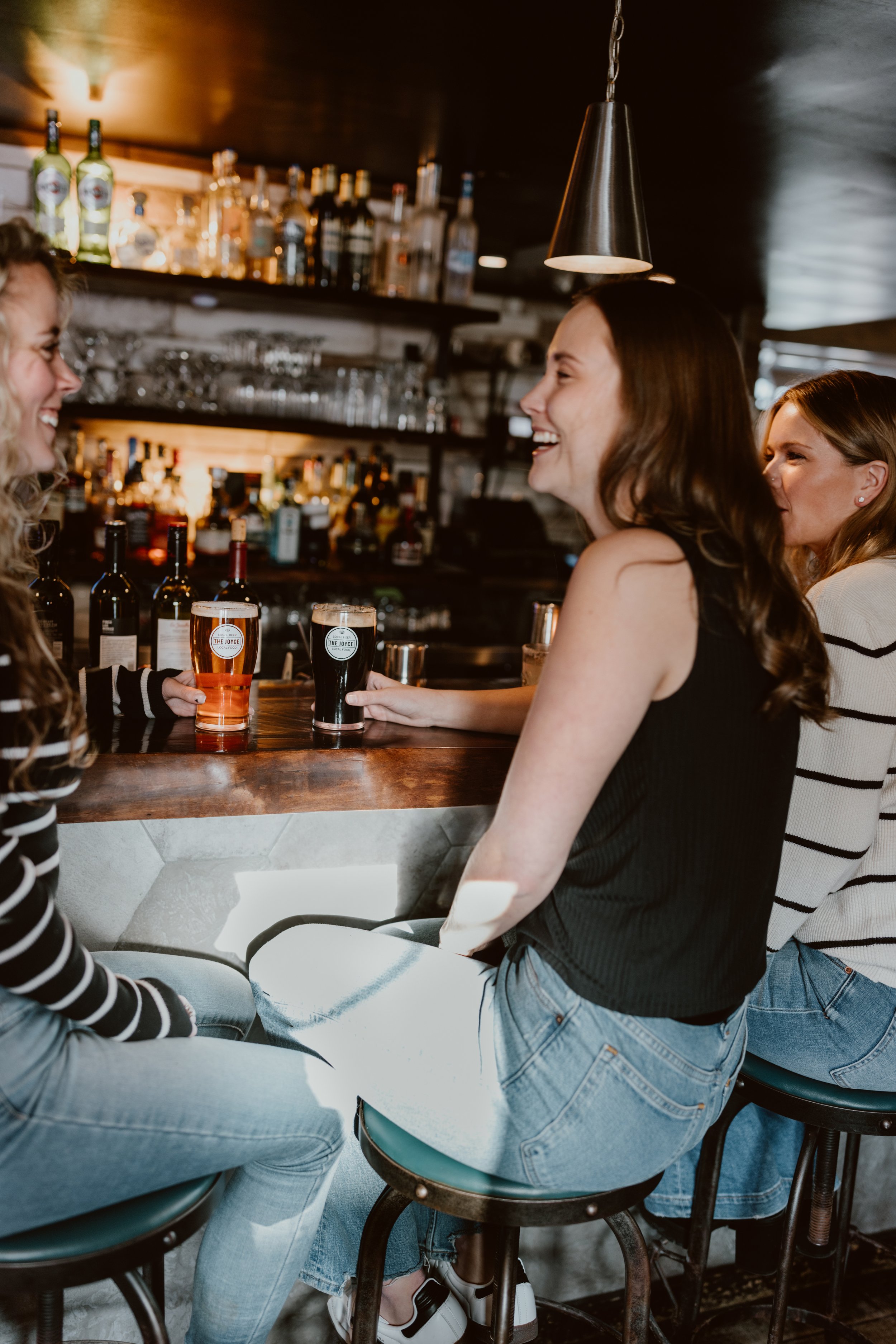Three women sitting at The Joyce bar, two with drinks at the bar counter, smiling and chatting with each other.