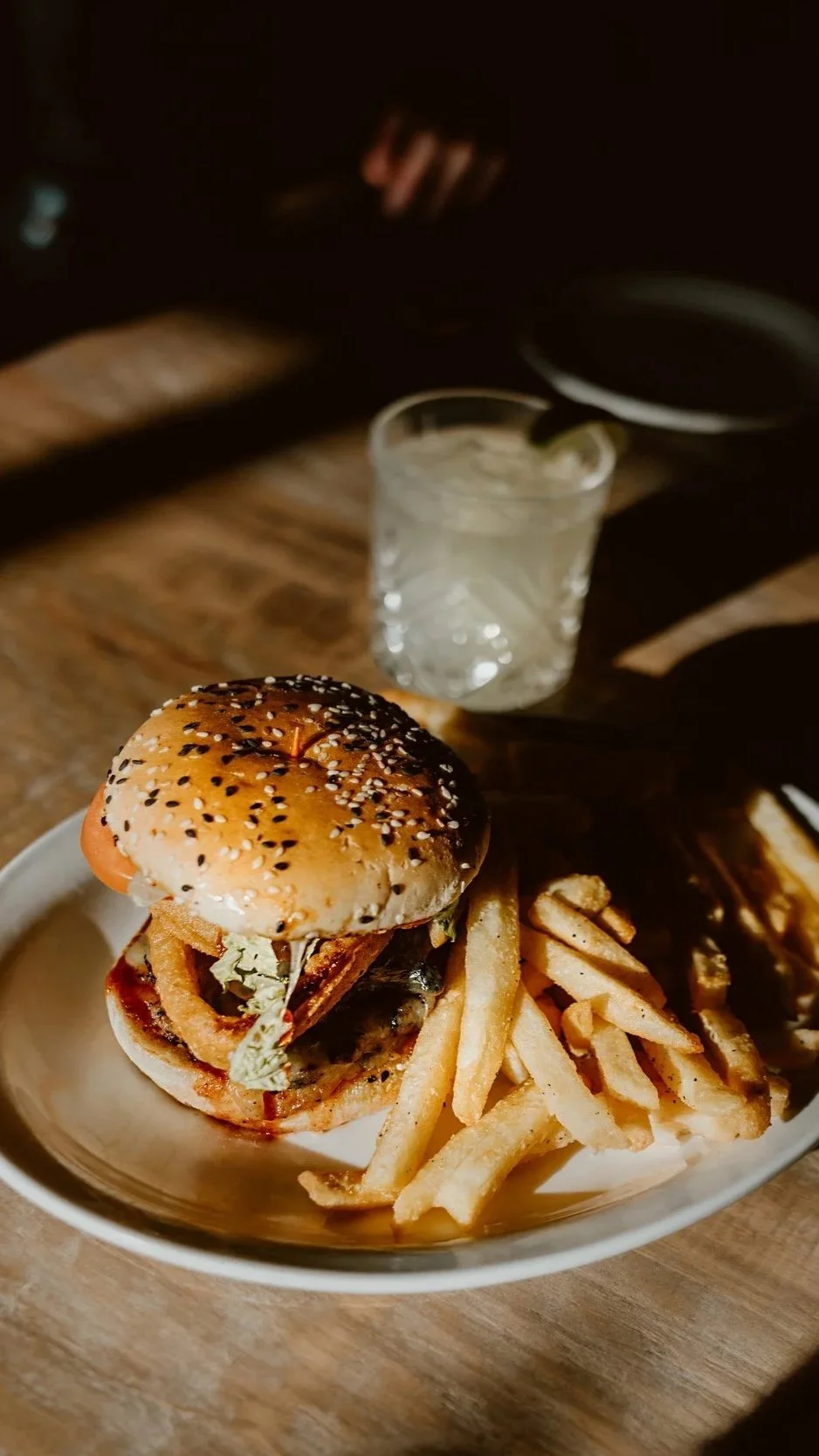 A plate with a cheeseburger topped with sesame seeds, onions, lettuce, tomato, and cheese, served with French fries, and a cocktail in the background at The Joyce Pub.
