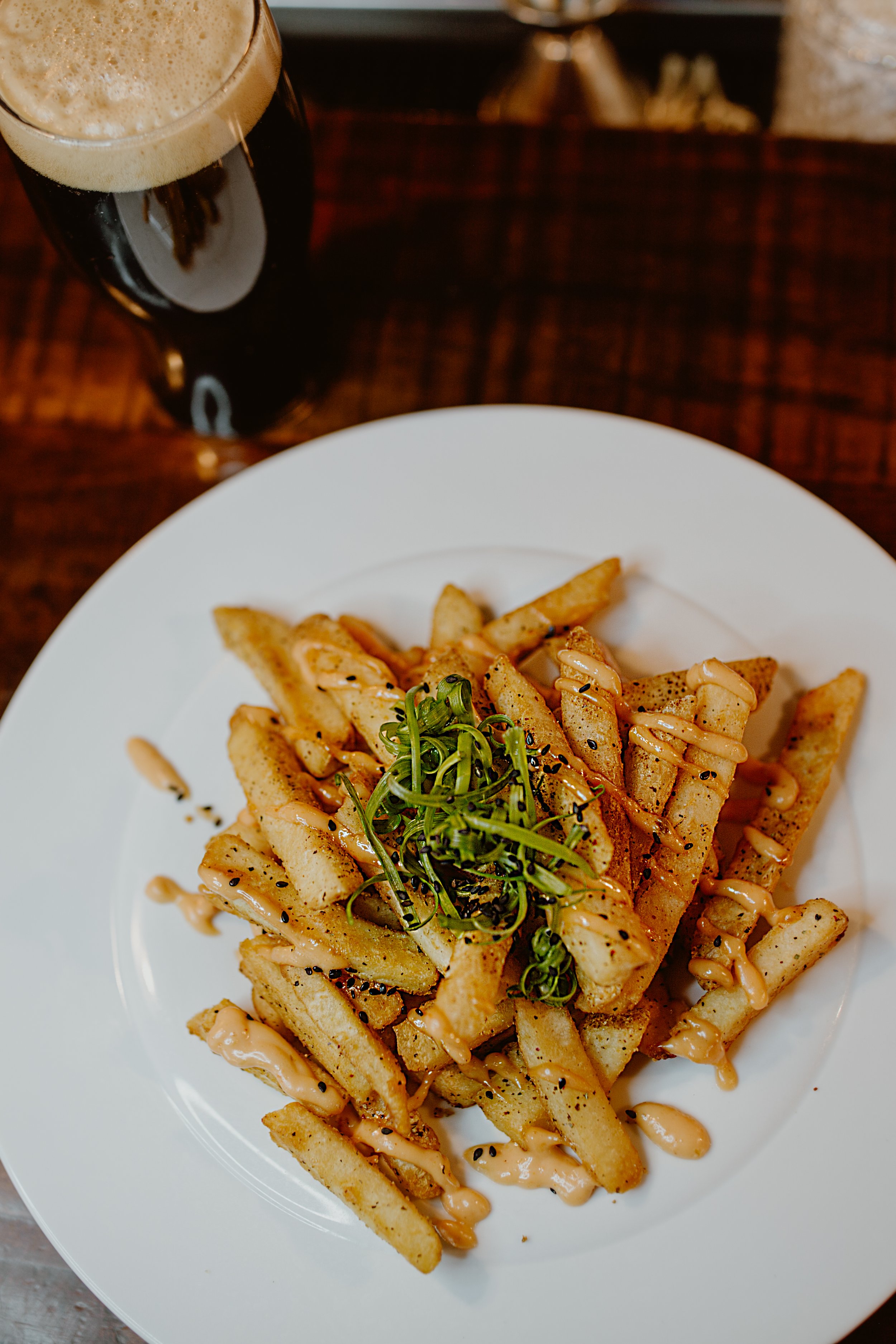 French fries topped with green onions, drizzled with creamy sauce, served on a white plate, with a craft beer in a glass at The Joyce pub.