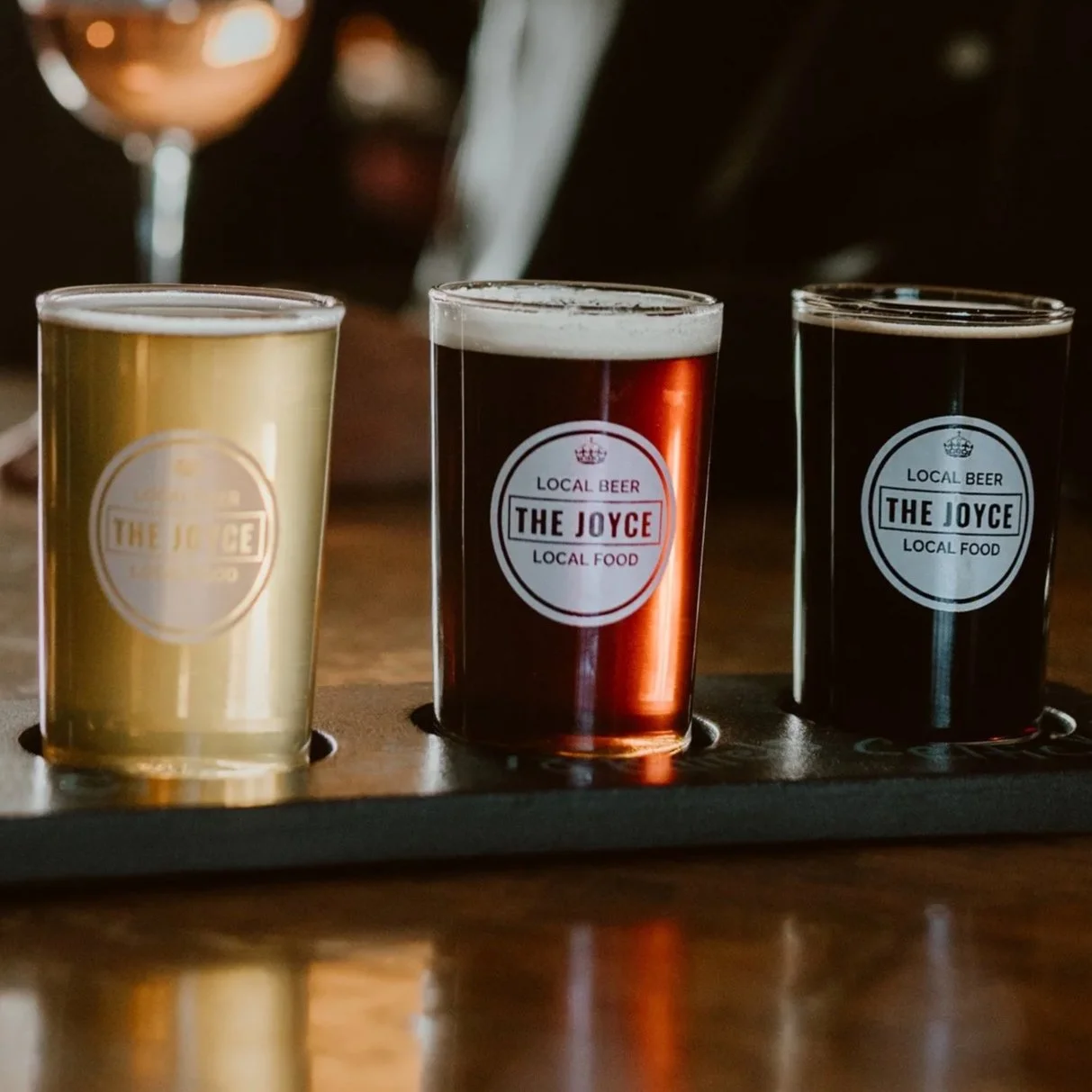 Three glasses of beer on a flight tray, with labels reading 'The Joyce' and descriptions of local beer and food, in a dimly lit bar setting.