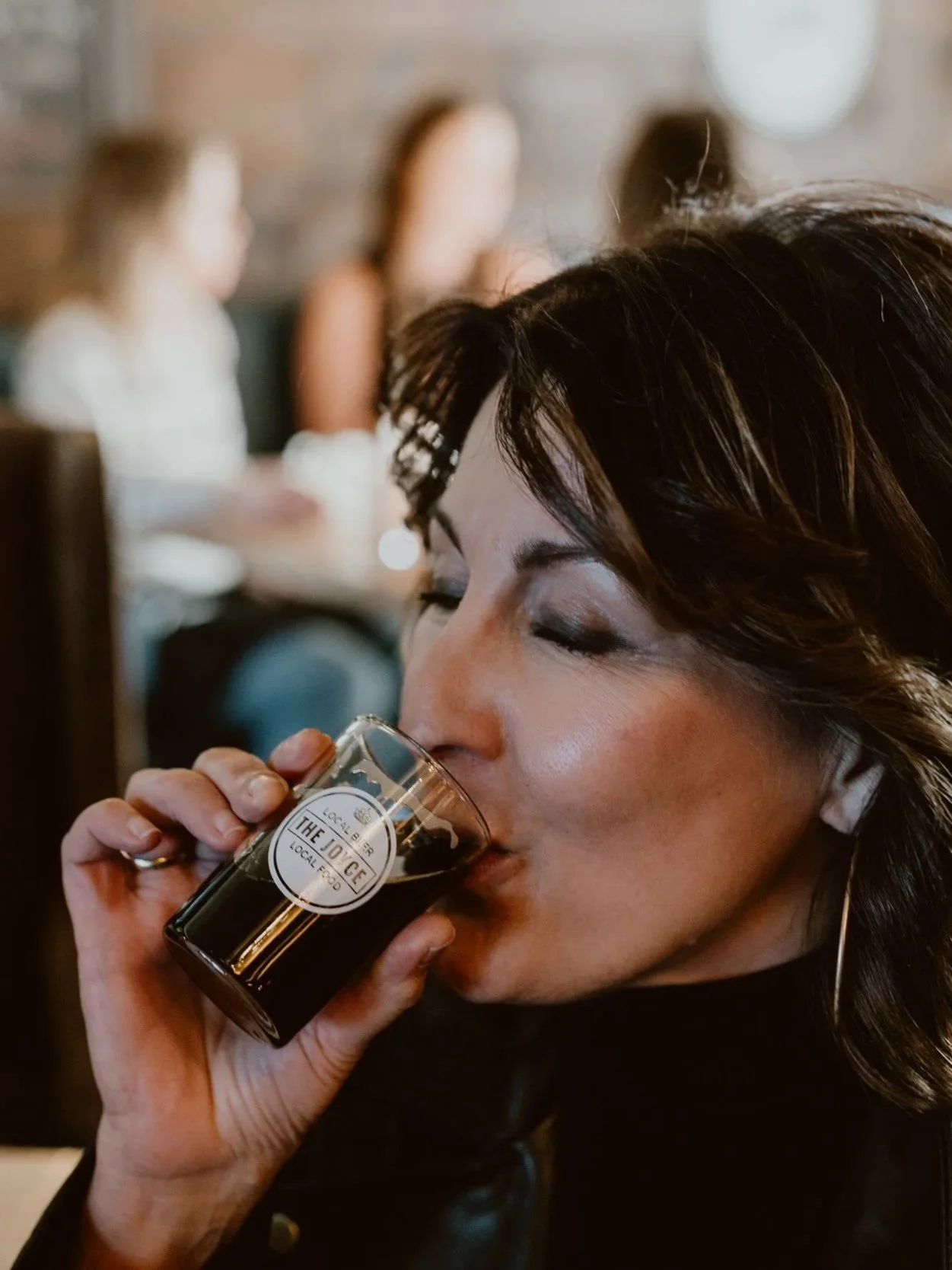 Woman drinking a dark craft beer from a glass with a label that reads "The Joyce" in The Joyce Pub with blurred background of other people.
