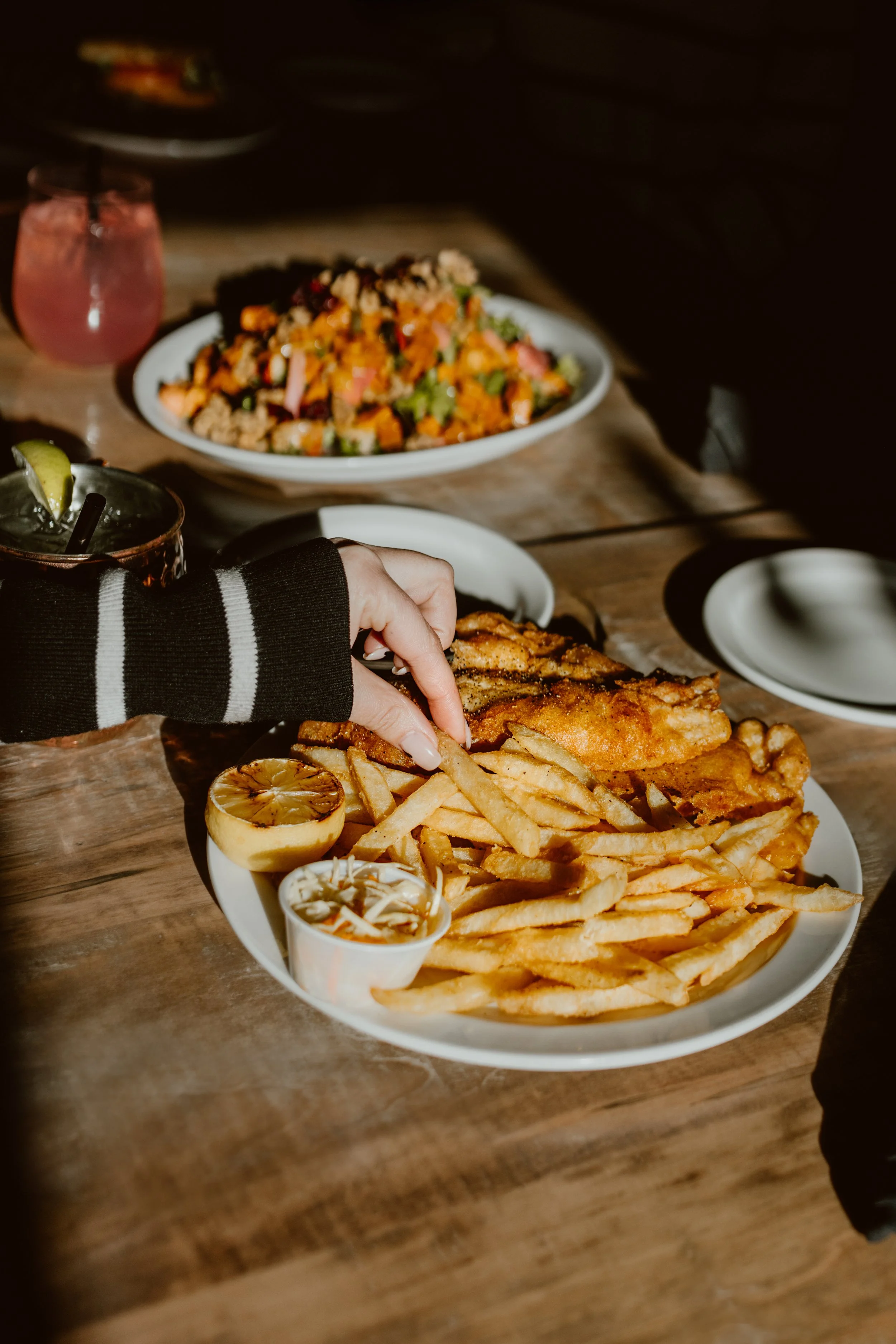 Person reaching for French fries and fried fish on a white plate, with lemon and condiment in small cups, on a rustic wooden table, with other plates and cocktails in the background at The Joyce Pub.