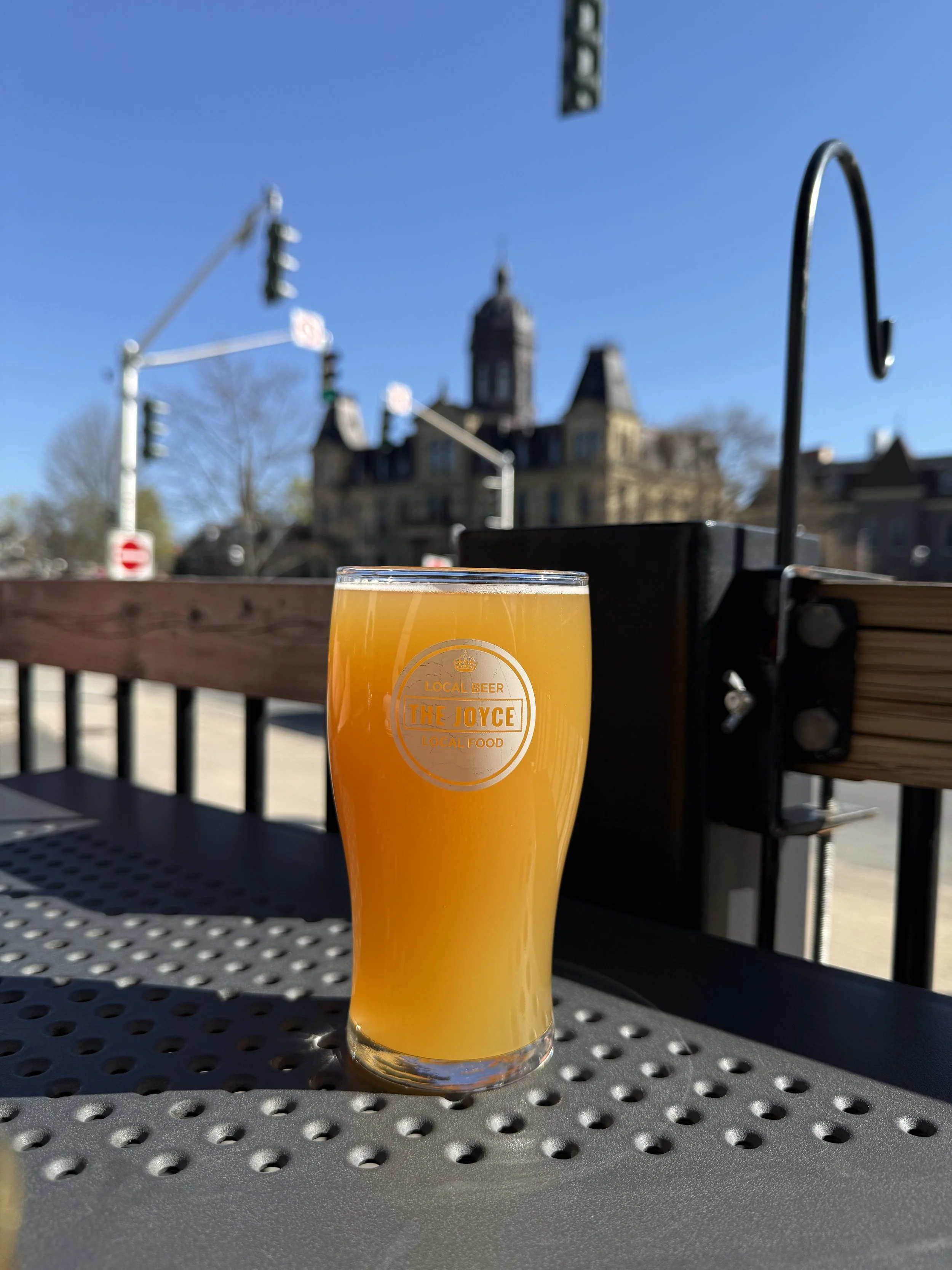 A pint glass filled with orange-coloured craft beer sitting on a perforated metal table outdoors. The glass has a logo that reads 'The Joyce, Local Beer, Local Food'. In the background, there's a historic building, under a clear blue sky.