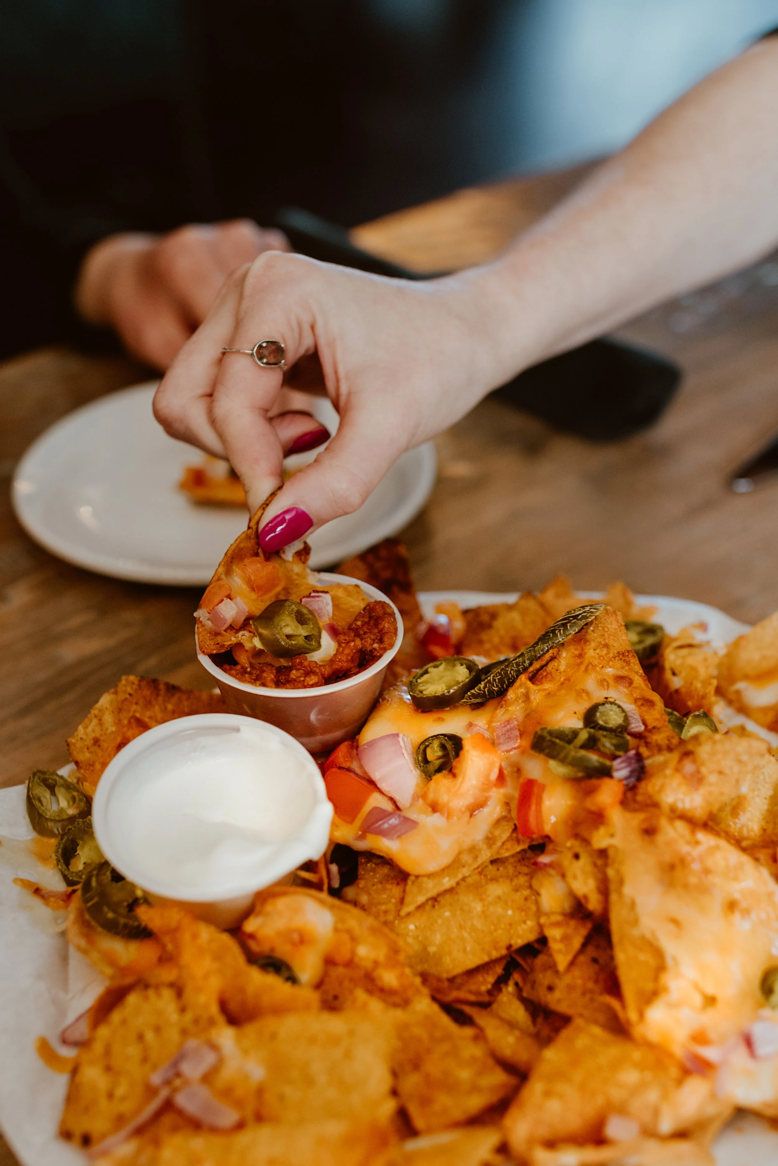 Close-up of a person's hand picking a slice of loaded nachos with jalapenos, onions, and cheese, on a tray with cheese dip and sour cream at The Joyce Pub.