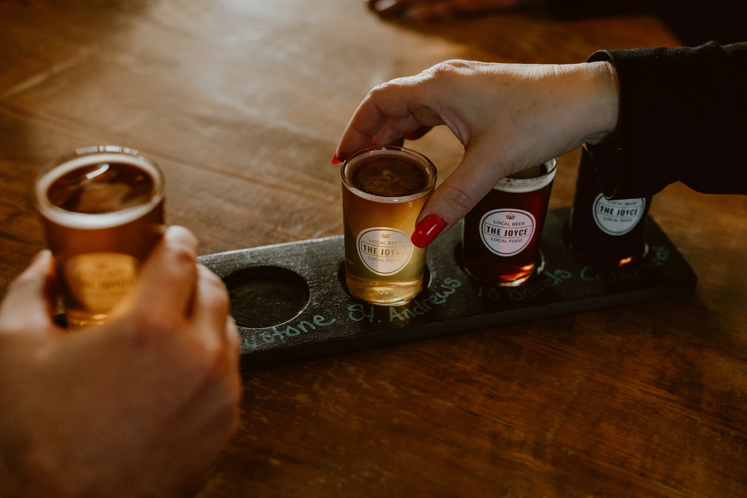 Two people holding small glasses of craft beer, with three glasses of craft beer on a tasting flight on a wooden table at The Joyce Pub.