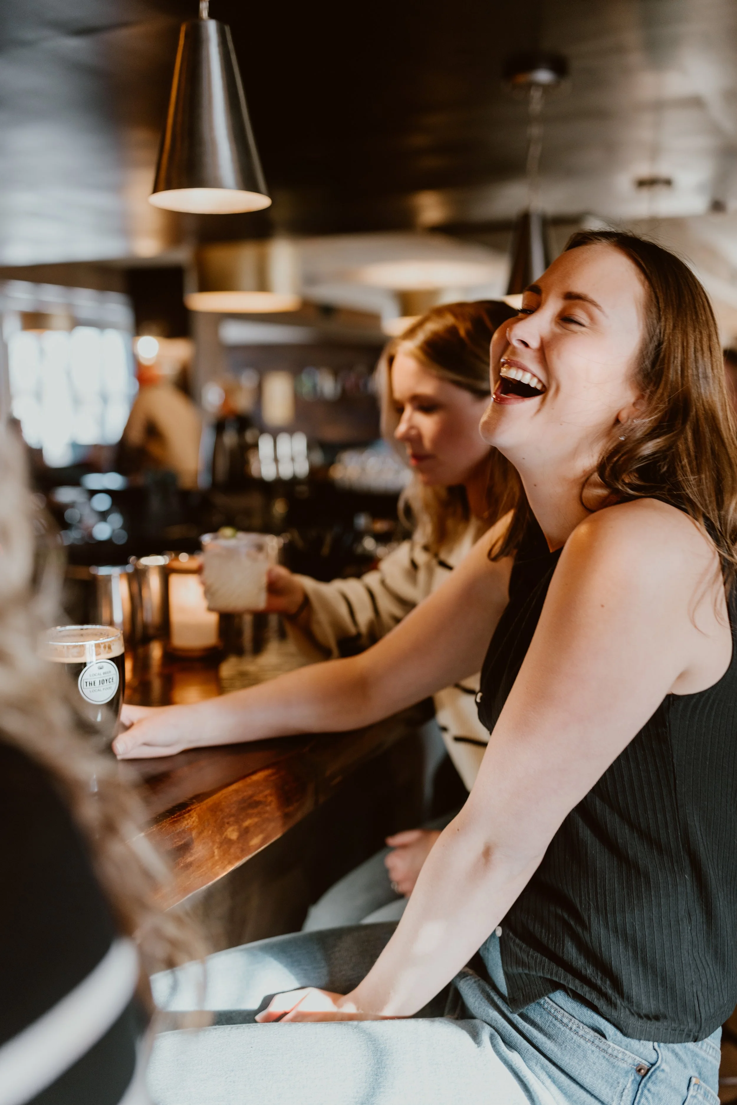 A woman is laughing with her mouth open at the bar of The Joyce Pub, sitting at the bar with other people, drink glasses of craft beer on the counter, warm lighting, and a dark ceiling.