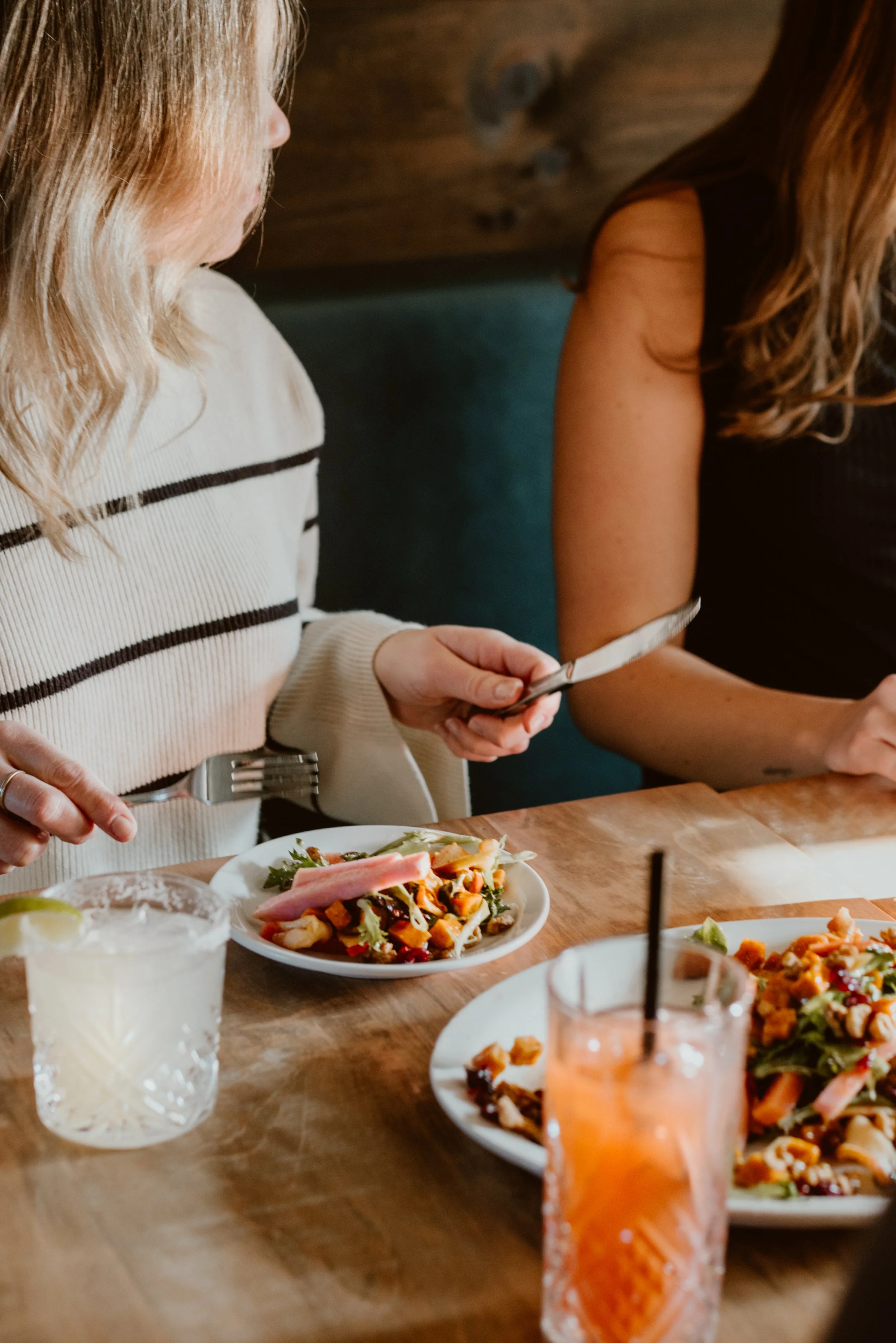 Two women sitting at a wooden table with salads and cocktails, one holding a knife, at The Joyce Pub.
