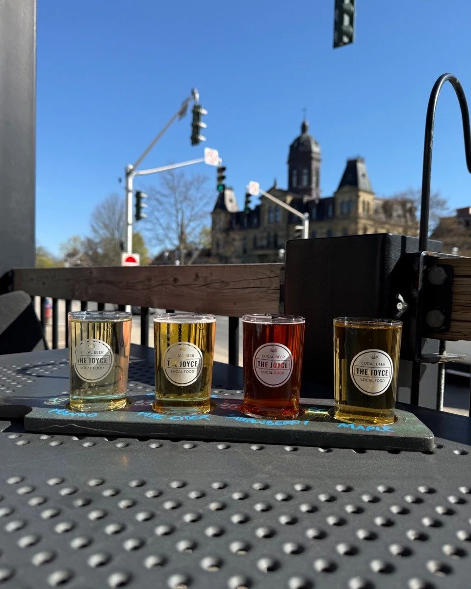 Four small glasses of craft beer from The Joyce Pub displayed on a flight paddle on an outdoor patio, with a city street and historic building visible in the background on a sunny day.