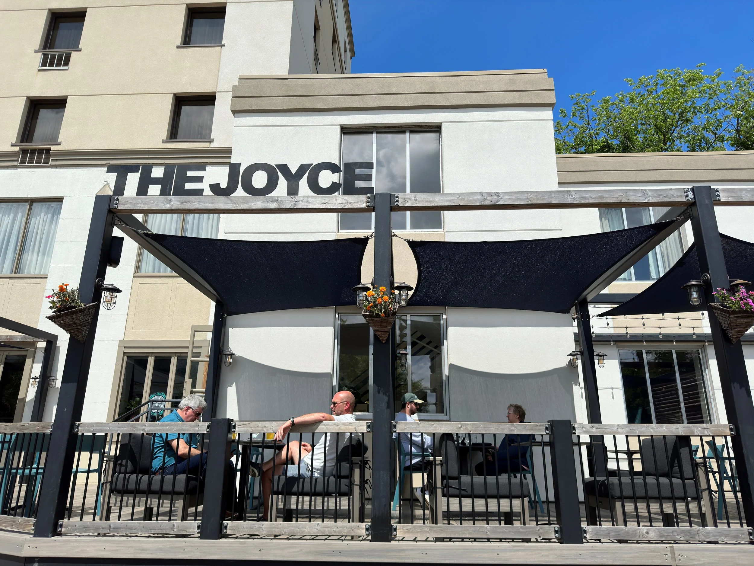 People sitting at an outdoor patio of a building named THE JOYCE, with black shade sails, planters with flowers, and string lights, under a blue sky.