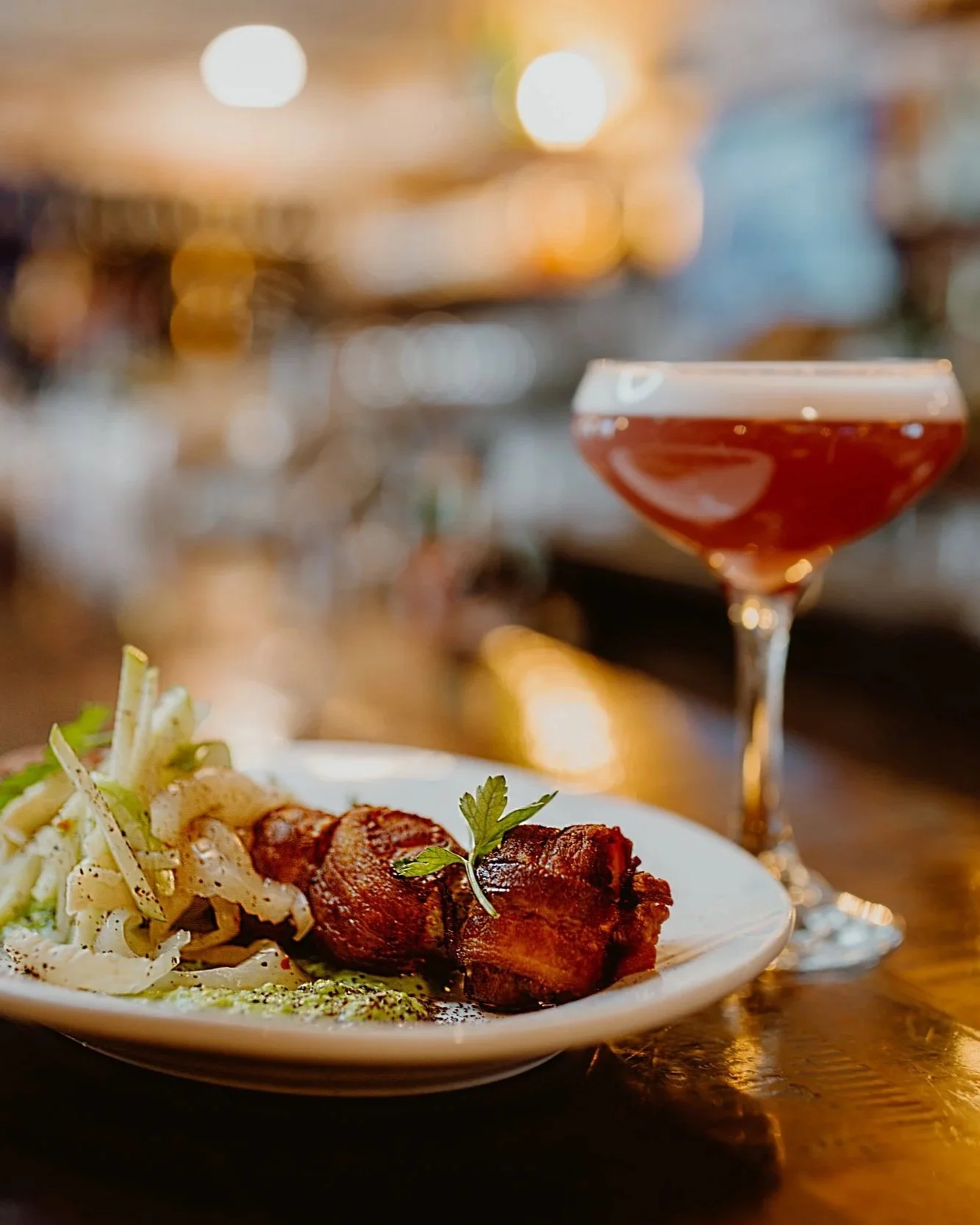 A plate of chicken wrapped scallops and a sprig of parsley, with cocktail on a wooden table in the dimly lit Joyce Pub.