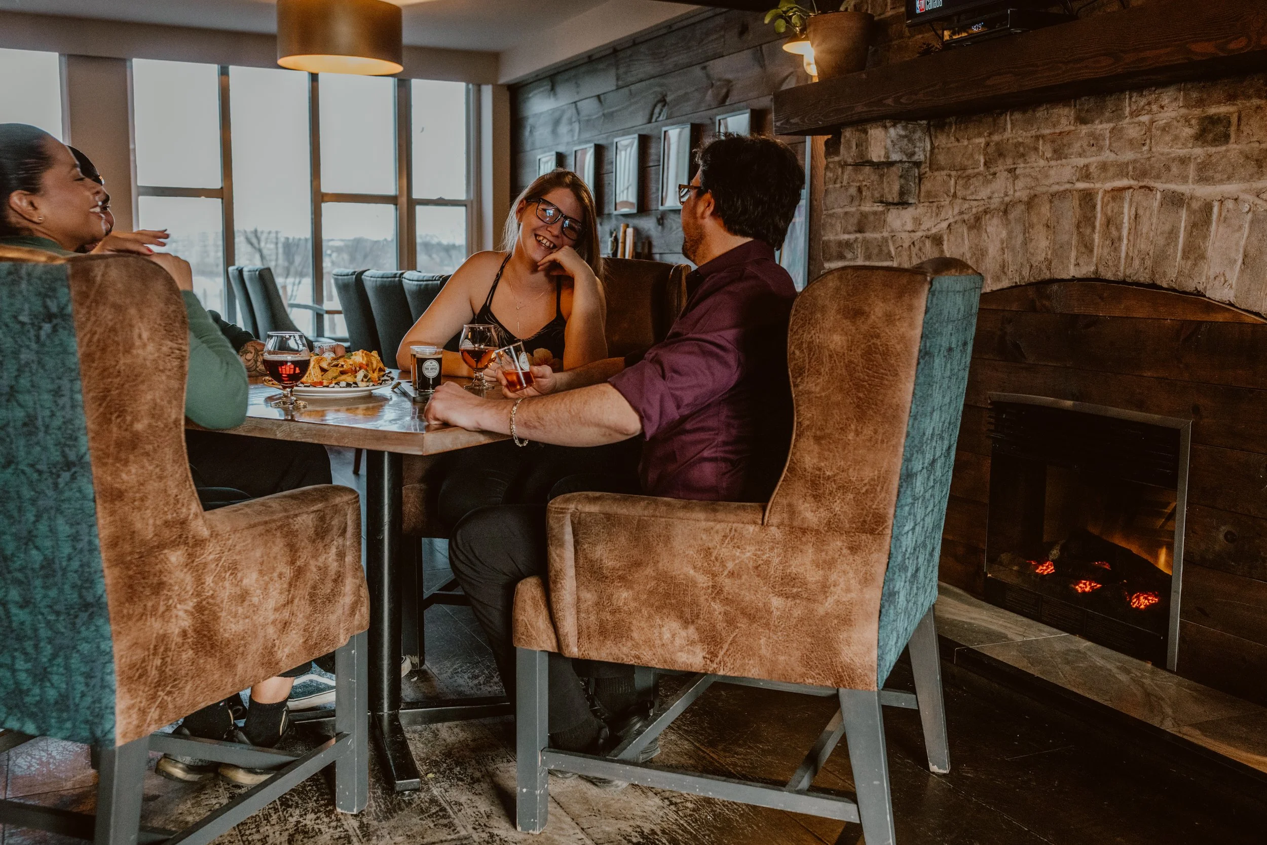 Four friends are sitting around a table in the cozy wingback chairs of The Joyce Pub, enjoying food and drinks. They are smiling and engaging in conversation. The pub has large windows, wooden walls, a fireplace, and warm lighting.