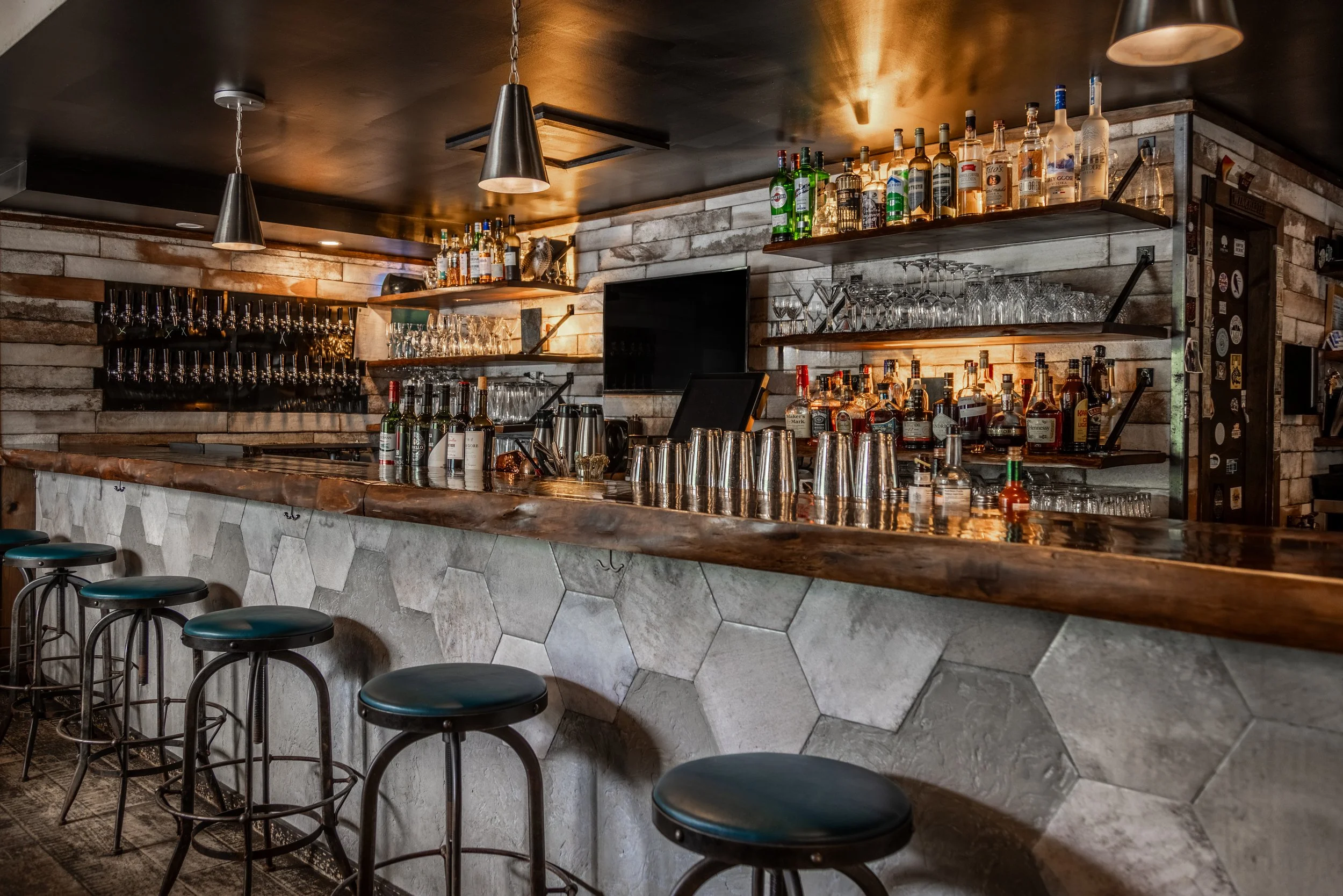 Empty bar with black stools, hexagonal tiles on the front, wooden countertop, shelves holding various liquor bottles and glassware, television, and pendant lights and the 36 craft beer taps of The Joyce Pub on the far wall.